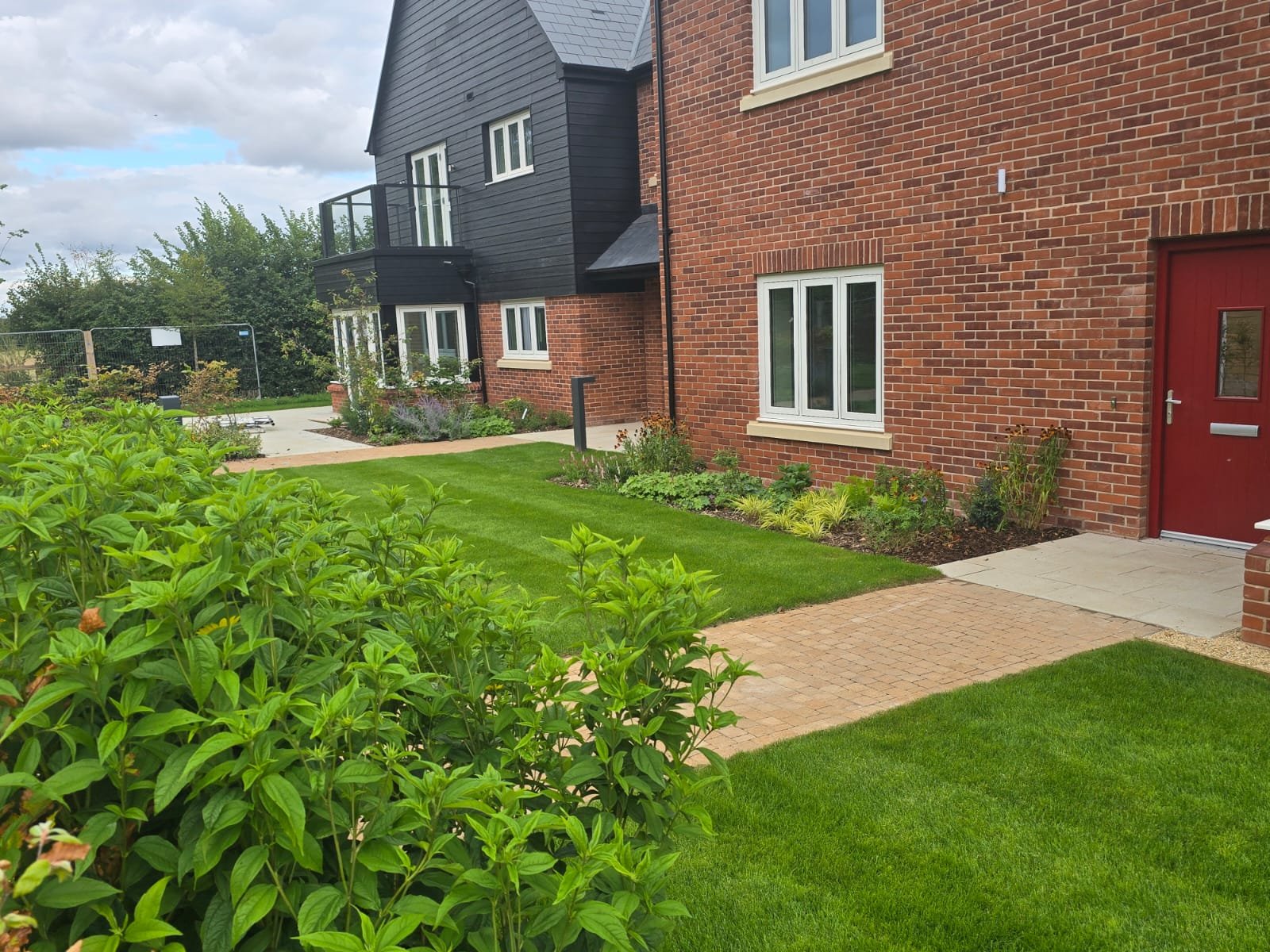 A well-maintained garden with green grass, flowering plants, and a brick and black exterior house with white windows and a red door.