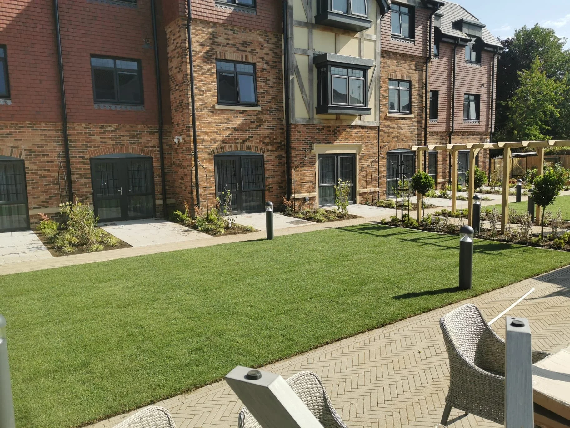 View of a residential building with brick and stucco exterior, multiple windows, small landscaped garden areas, well-maintained grass lawn, pathway, and outdoor seating.
