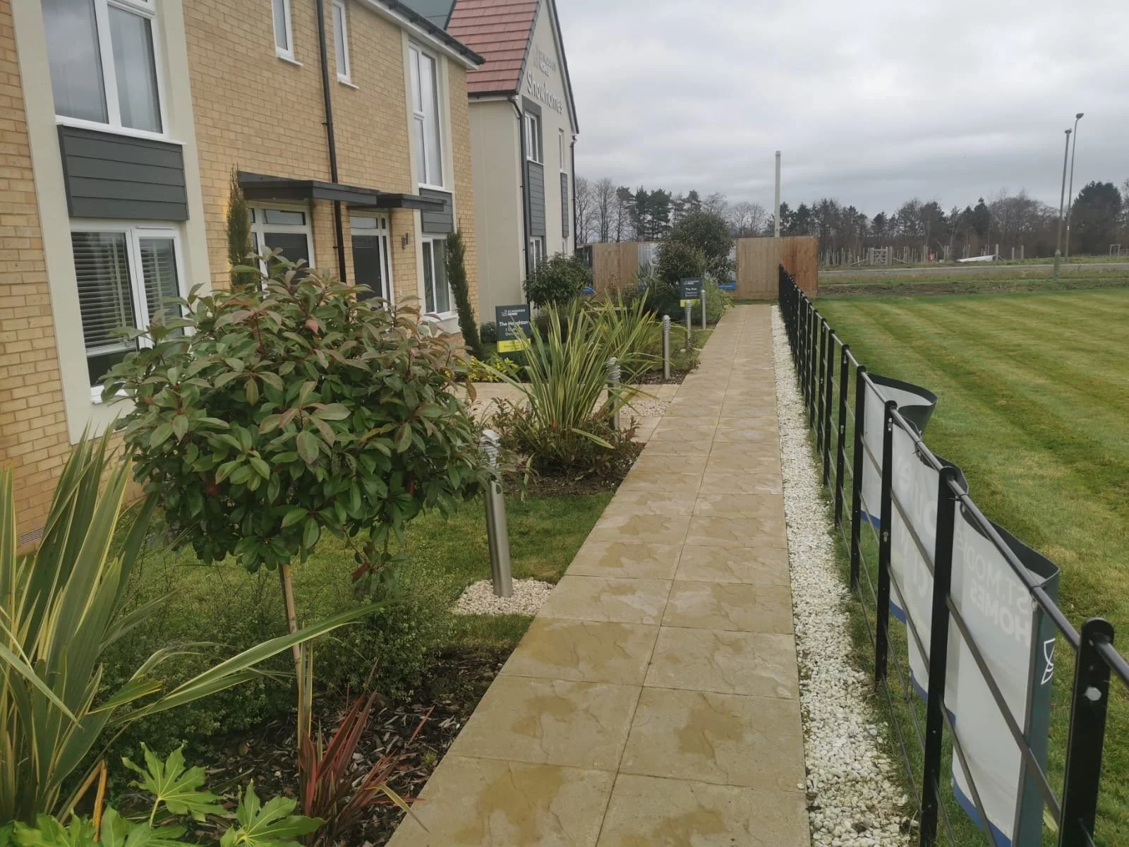A paved sidewalk next to a garden with bushes and plants, bordered by a black metal fence, with a grassy field to the right and residential buildings to the left, under an overcast sky.