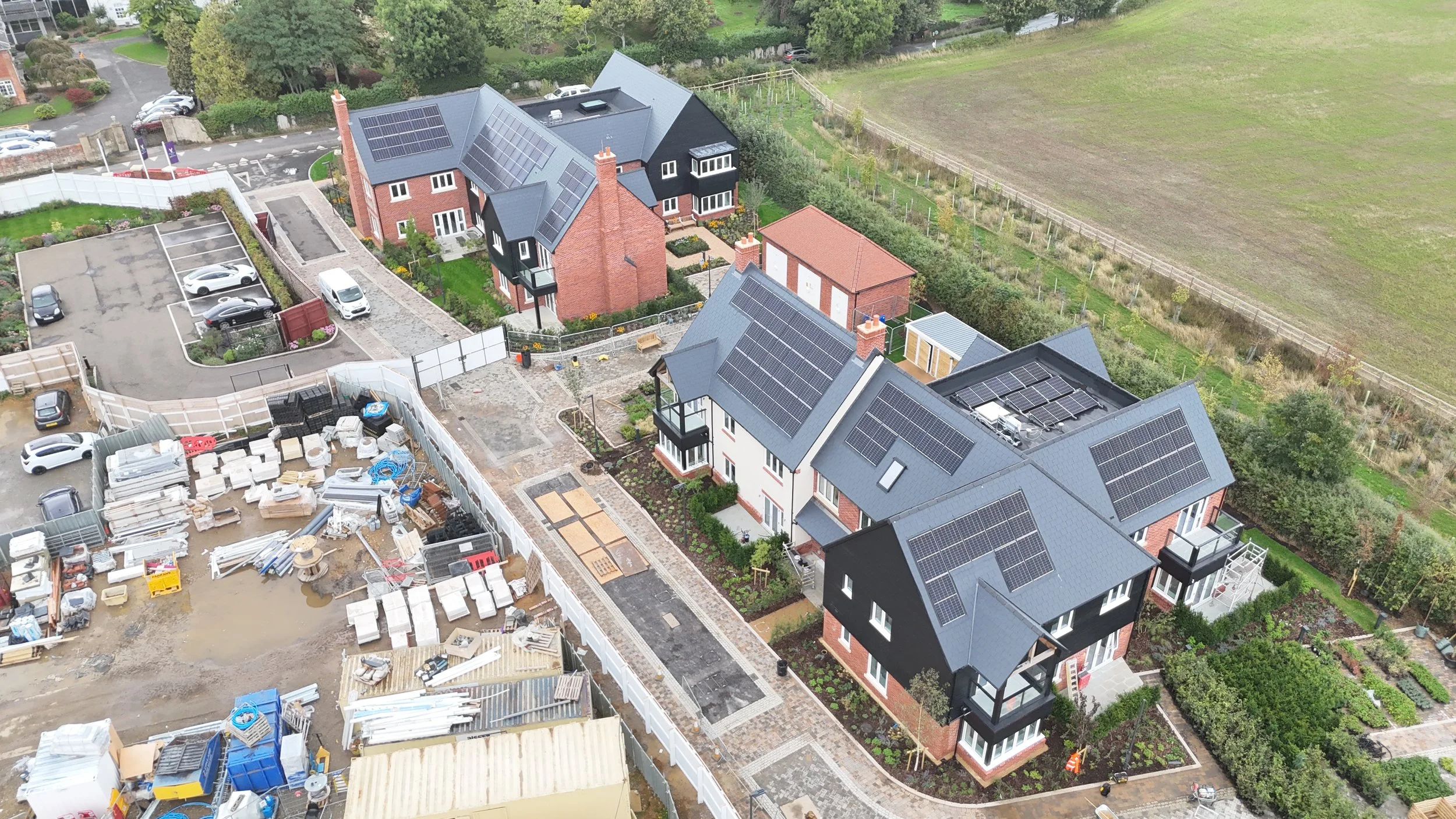 Aerial view of new residential buildings with solar panel roofs, surrounded by gardens and a construction site with building materials and equipment.