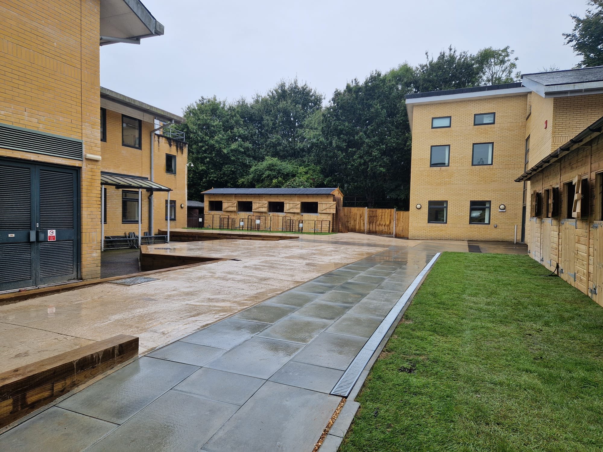 Empty courtyard with wet pavement, surrounded by yellow brick buildings and a small wooden shed, with trees in the background on a cloudy day.