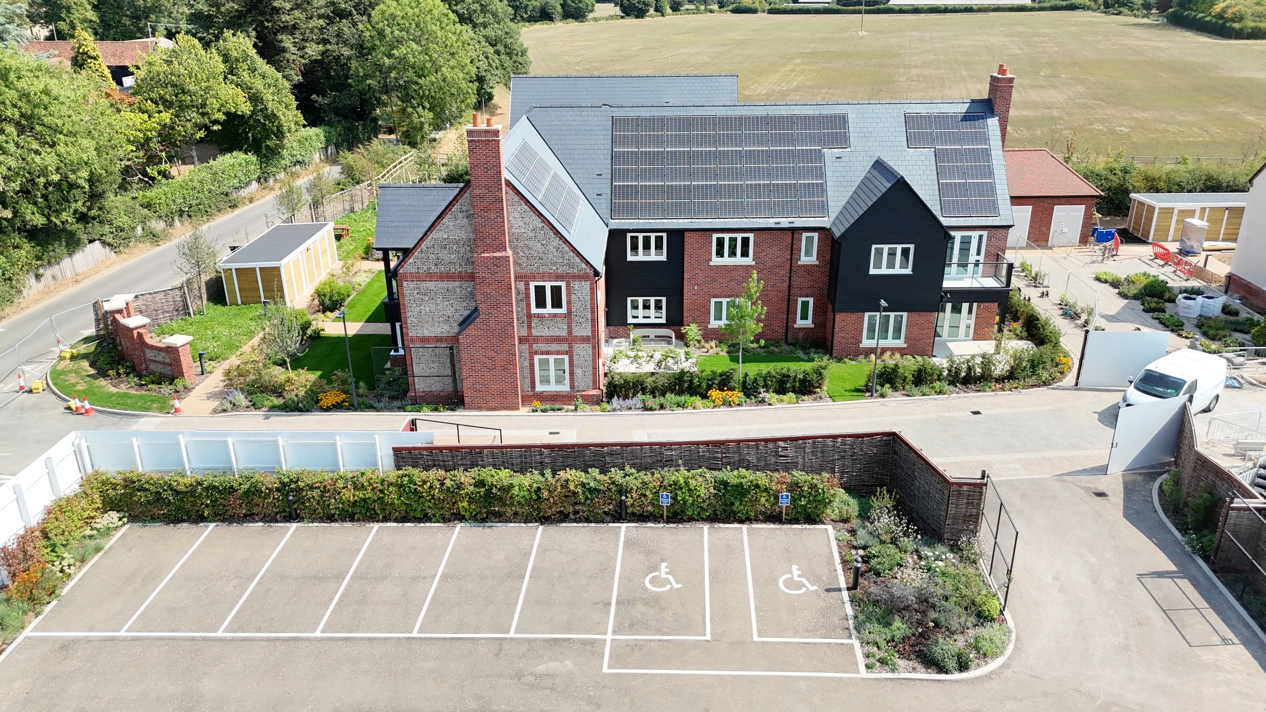 Aerial view of a modern brick residential building with solar panels on the roof, surrounded by a landscaped garden and parking lots with designated handicapped parking spaces.