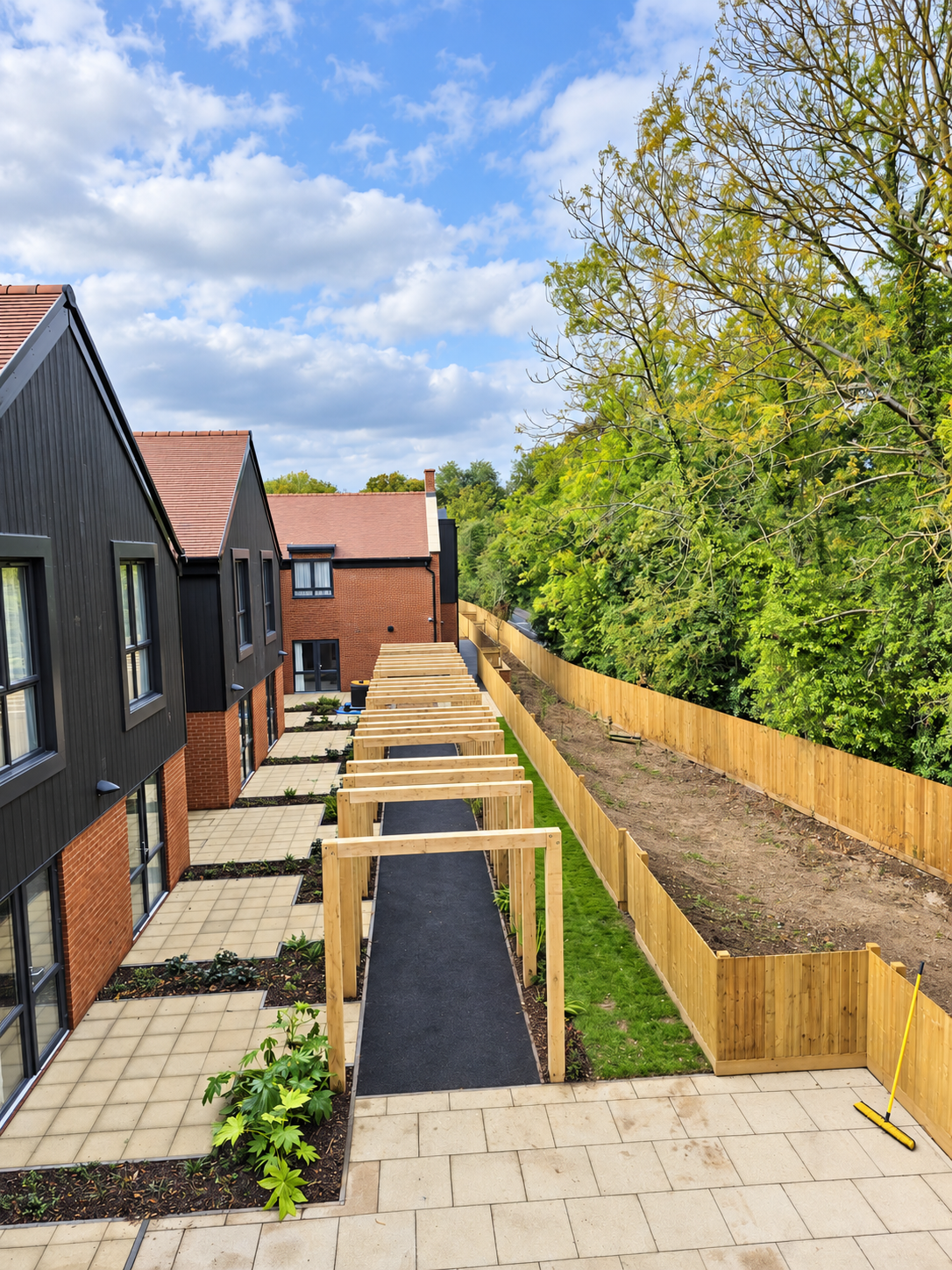 Newly constructed backyard with wooden framework for future patio or canopy, paved walkways, and a fenced garden area adjacent to modern black and brick townhouses under a partly cloudy sky.