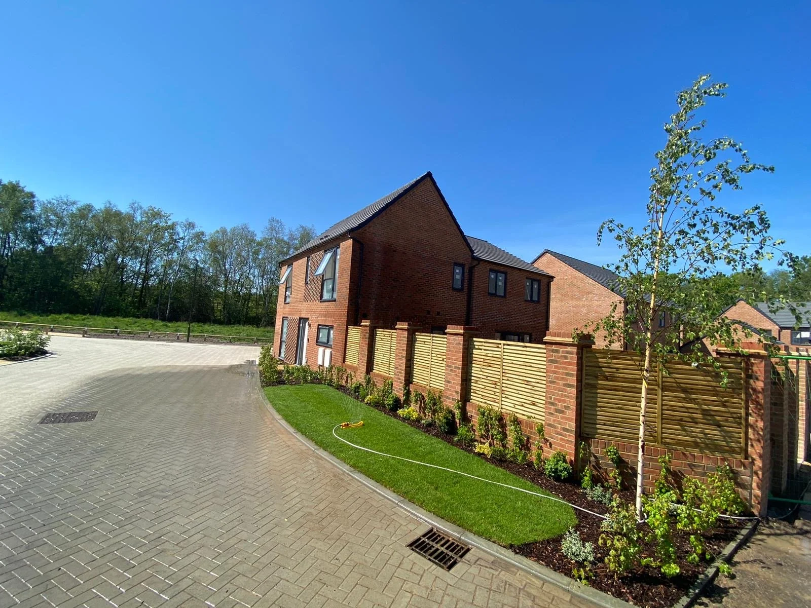 Newly built brick residential houses with modern design, surrounded by landscaped gardens, a paved driveway, and a wooden fence on a clear sunny day.