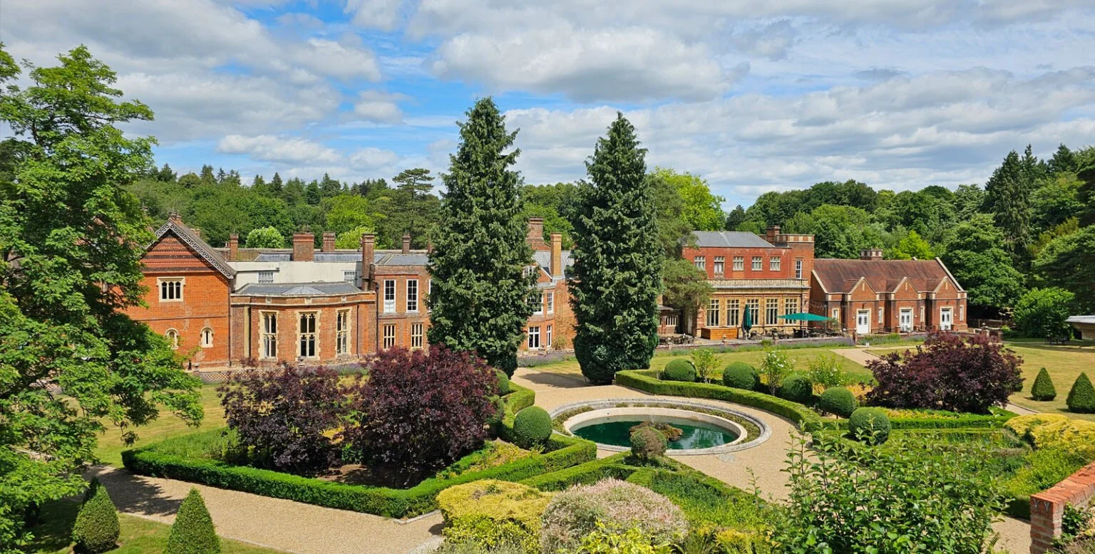 Beautiful landscaped garden with trimmed bushes and a circular pond, in front of a large brick mansion surrounded by lush trees and a partly cloudy sky.