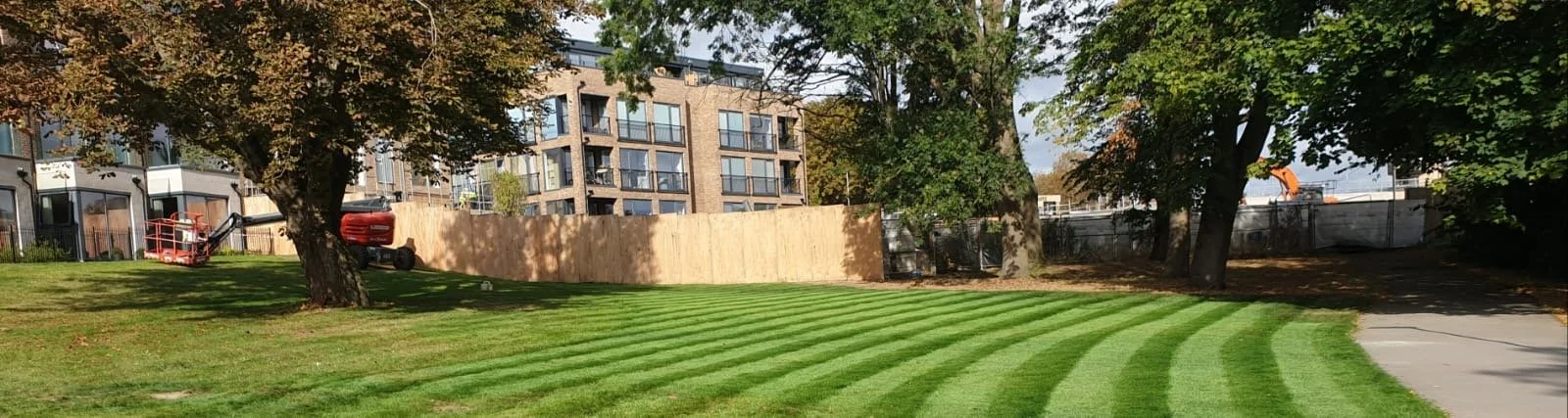 A grassy park with fresh mowed stripes, surrounded by trees, with apartment buildings and a construction site in the background.