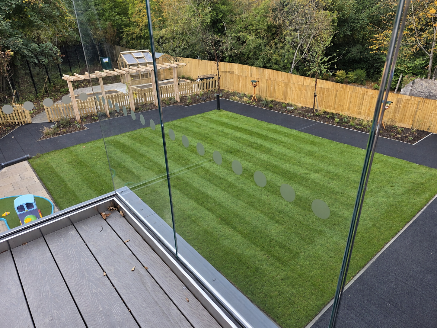 View from a balcony overlooking a backyard with a green grass lawn surrounded by a black border, a wooden fence, and trees, with a small play area and garden beds.