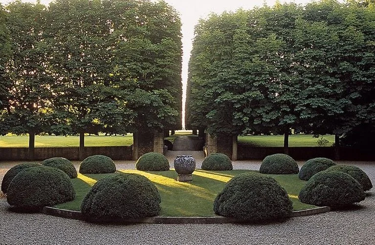 A symmetrical garden with neatly trimmed bushes, a central decorative urn, large trees framing a pathway, and a gravel ground.