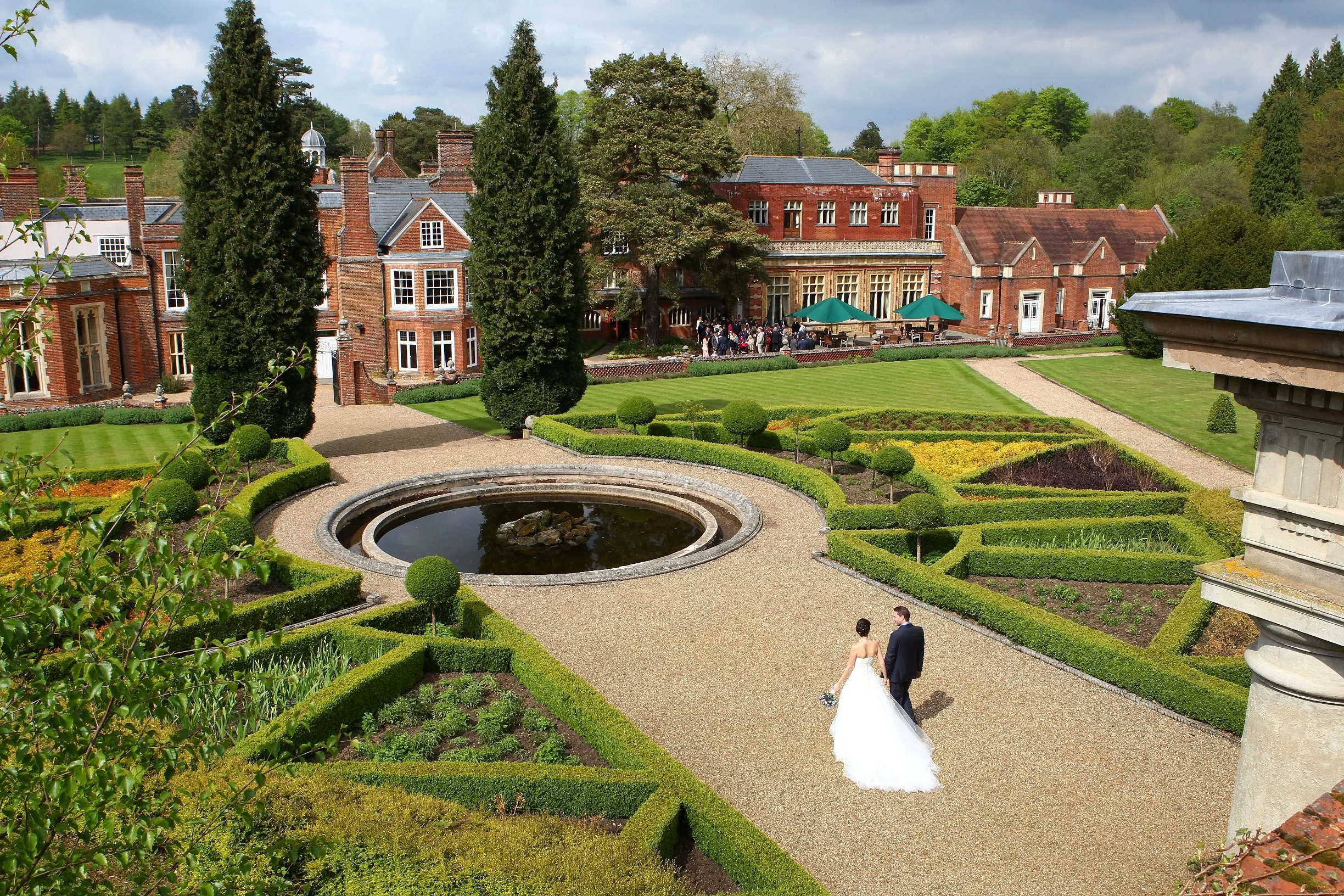 A bride and groom walking through a manicured garden with a fountain, surrounded by historic red-brick buildings, on a cloudy day.