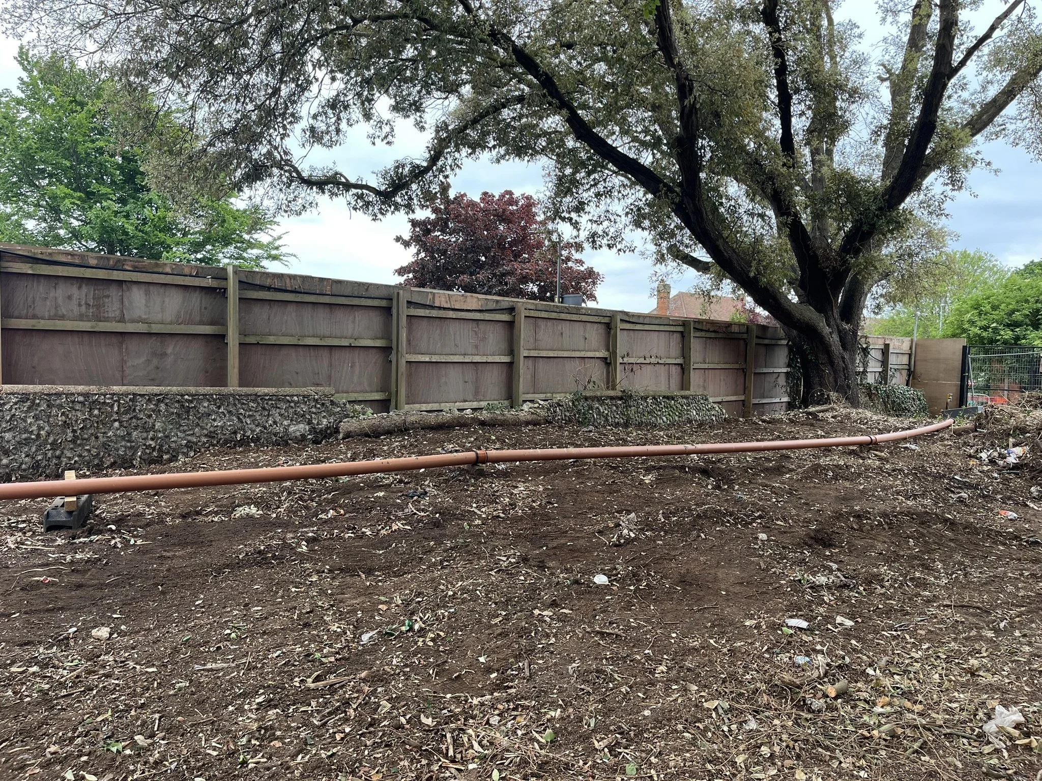 A backyard with dug-up dirt, a large tree, and a brown wooden fence. There is a copper pipe laid out on the ground. The sky is partly cloudy.