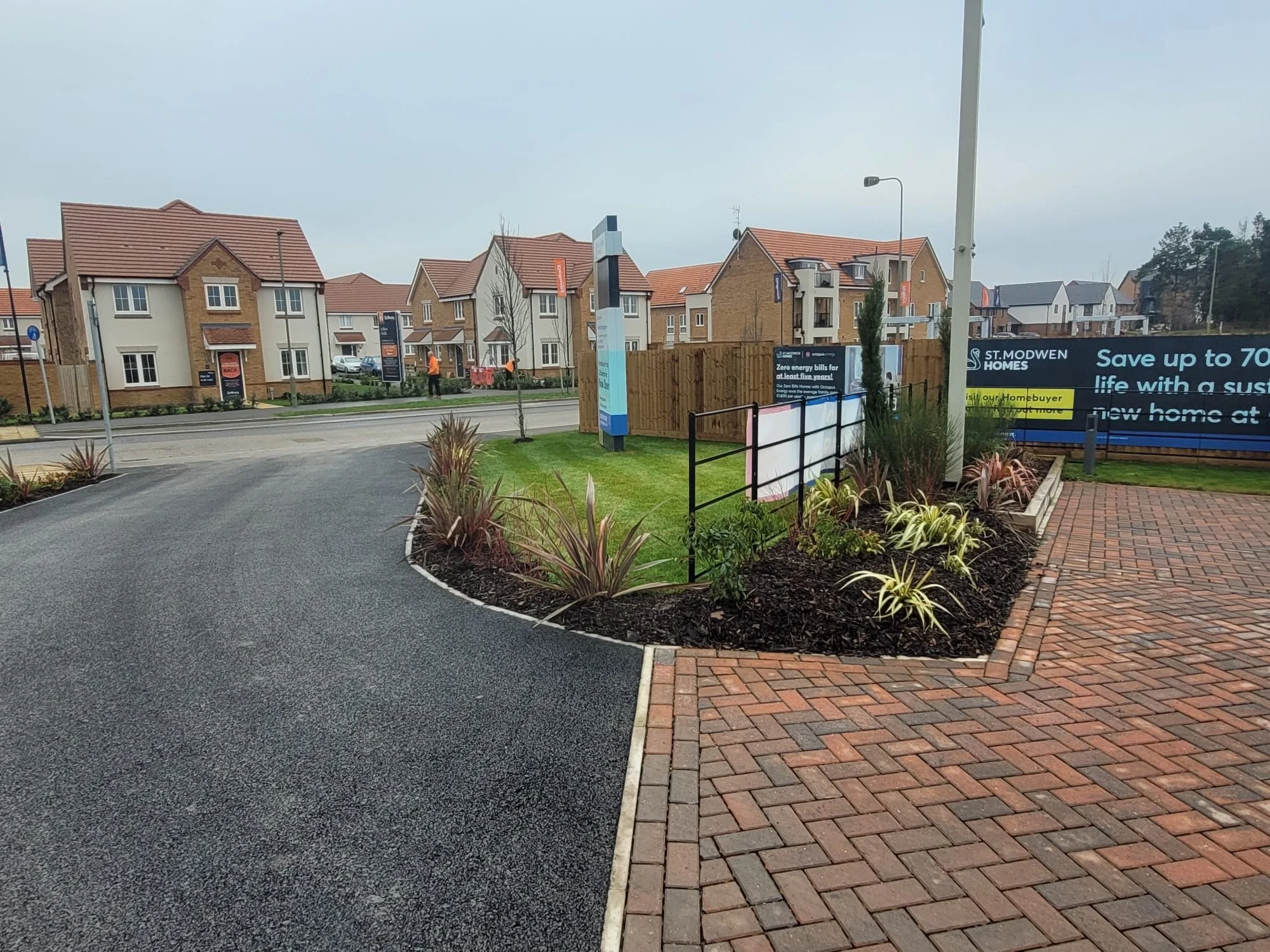 Residential area with modern multi-story houses with pitched roofs, a landscaped foreground with various plants, a paved pathway, and construction signs.