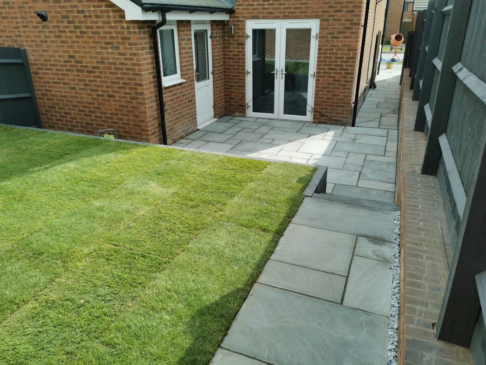Backyard with a grassy lawn and a paved patio area next to a brick house with white-framed glass doors.