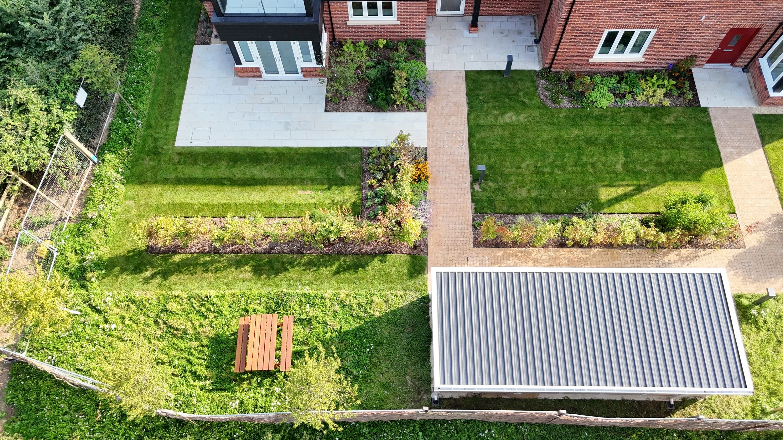 Aerial view of a residential backyard with green lawn, flower beds, paved walkways, a shed with a metal roof, and a small wooden bench surrounded by trees and bushes.