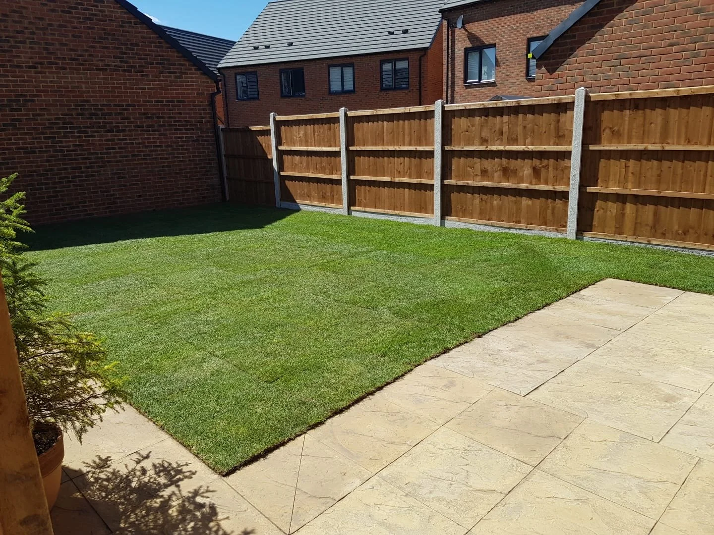 A backyard with a well-maintained green lawn, a stone patio area, and a wooden fence. There are brick houses in the background and a small potted plant in the corner.