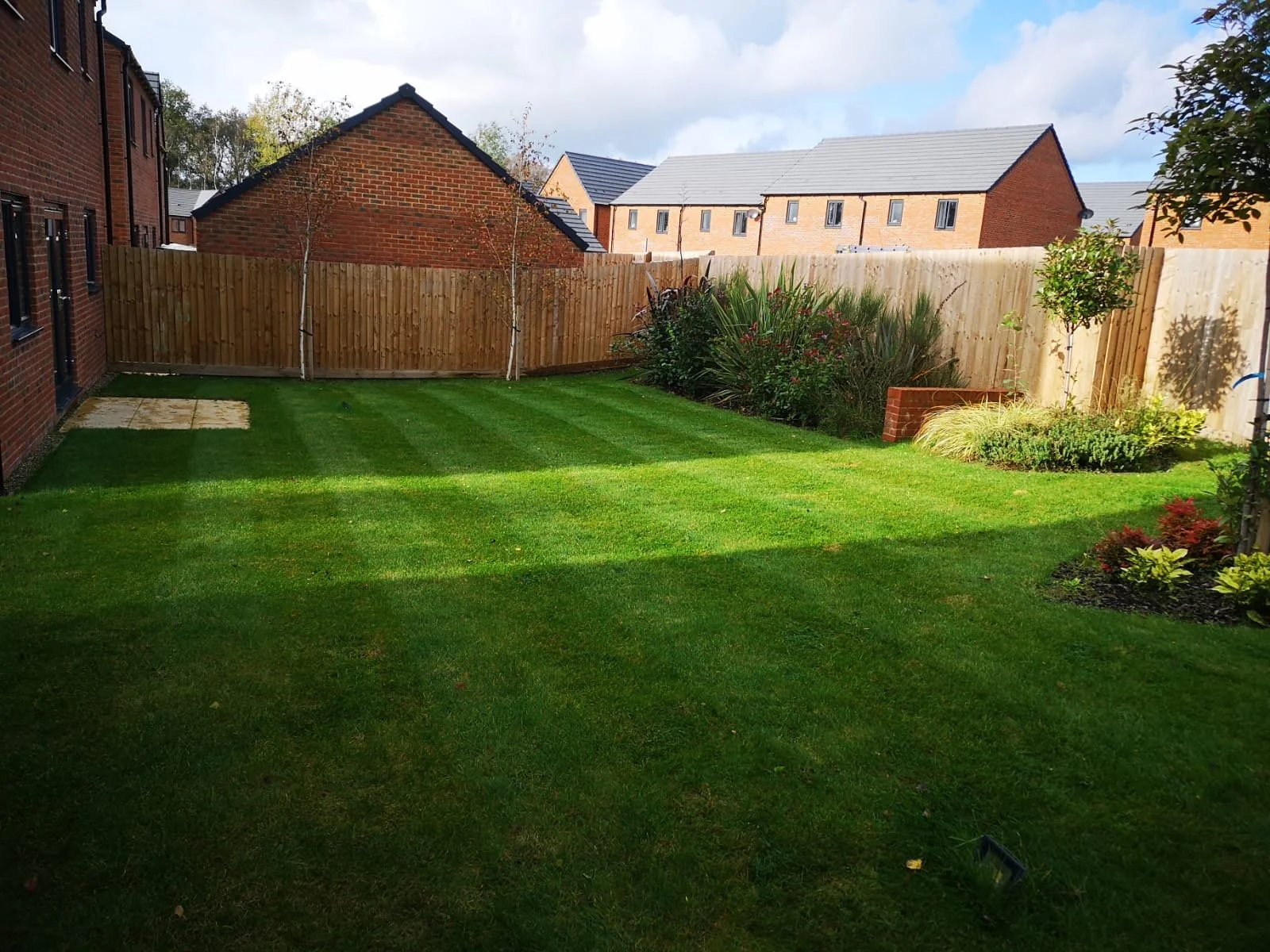 Clean, well-maintained backyard with striped green grass, small garden beds, and a wooden fence. There are neighboring brick houses and trees in the background, under a partly cloudy sky.