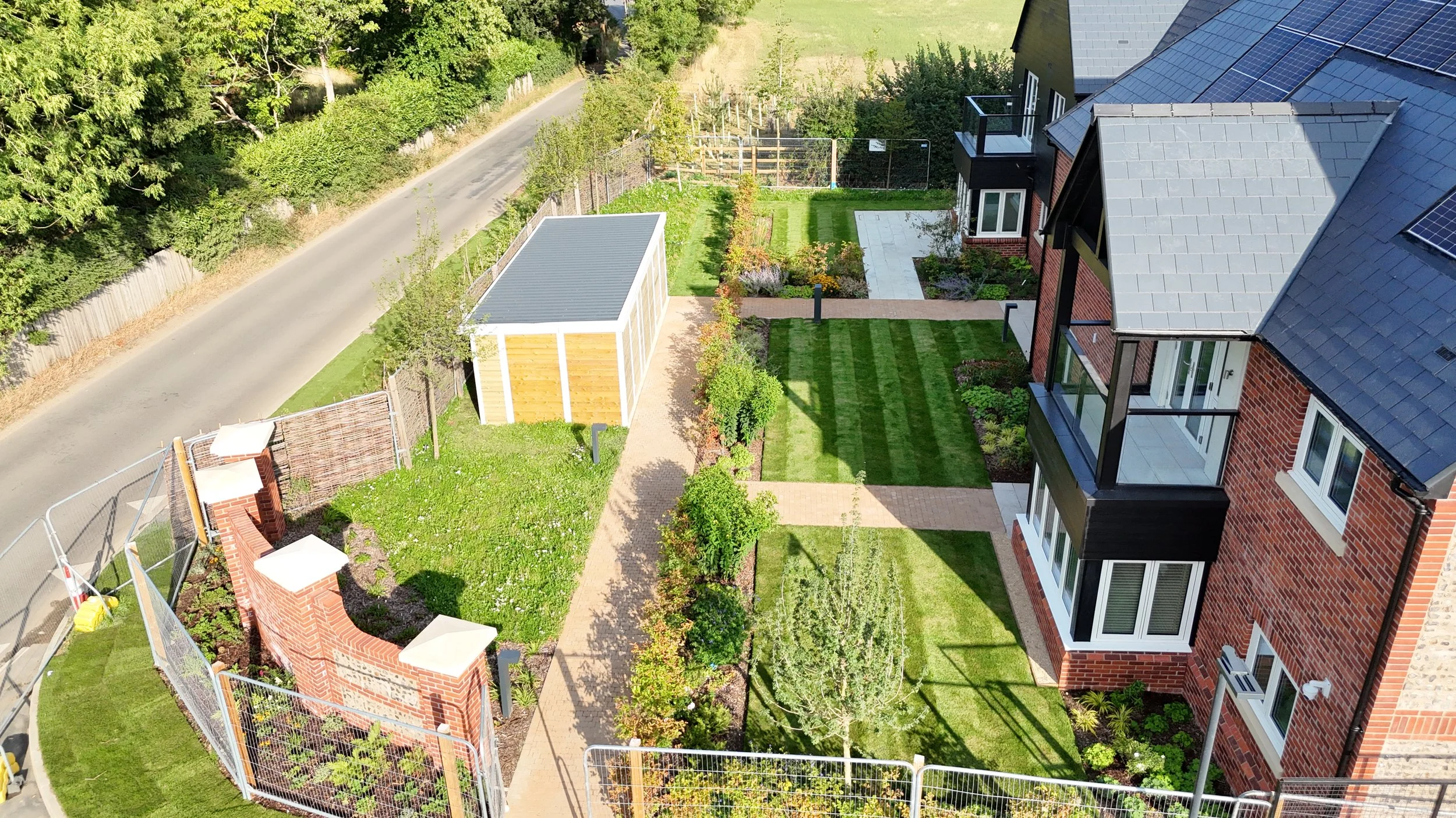 Aerial view of a modern residential garden with well-maintained lawns, pathways, small trees, and a brick building with balconies, adjacent to a street with a hedge and trees in the background.