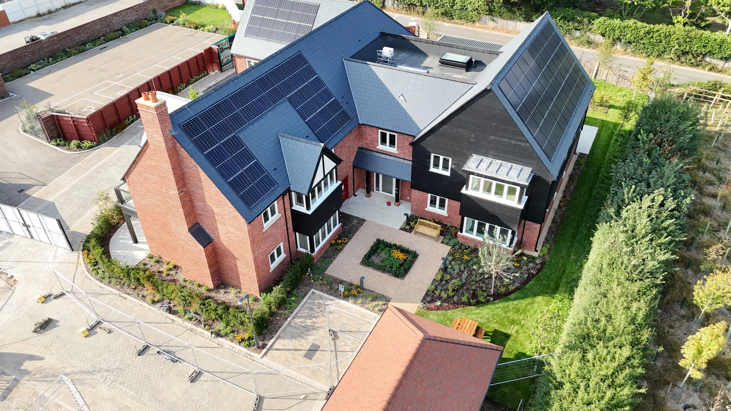 Aerial view of a modern brick house with solar panels on the roof, a small front garden with a pathway and a bench, surrounded by trees and neighboring buildings.