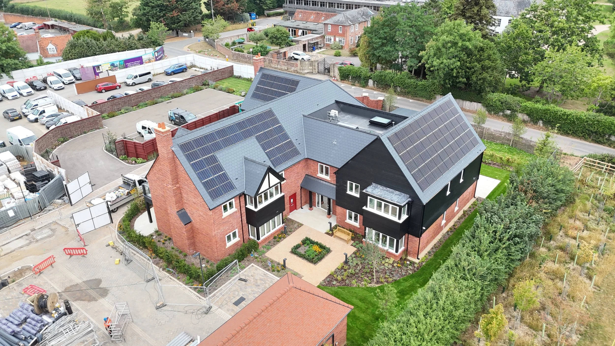 Aerial view of a modern residential building with solar panels on the roof, surrounded by a garden and neighboring houses, with a parking lot and construction area nearby.