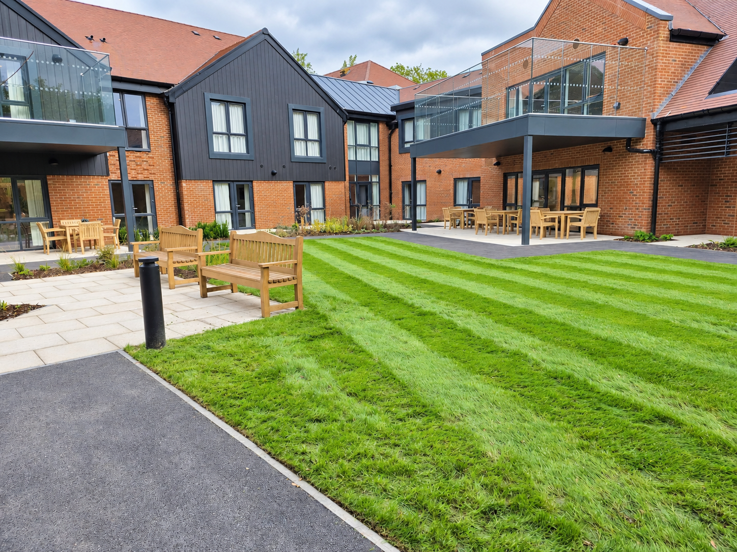 Modern residential apartment complex with brick and black wooden exterior, balconies with glass railings, neatly maintained lawn with striped grass, paved walkways, and wooden benches and outdoor dining tables.