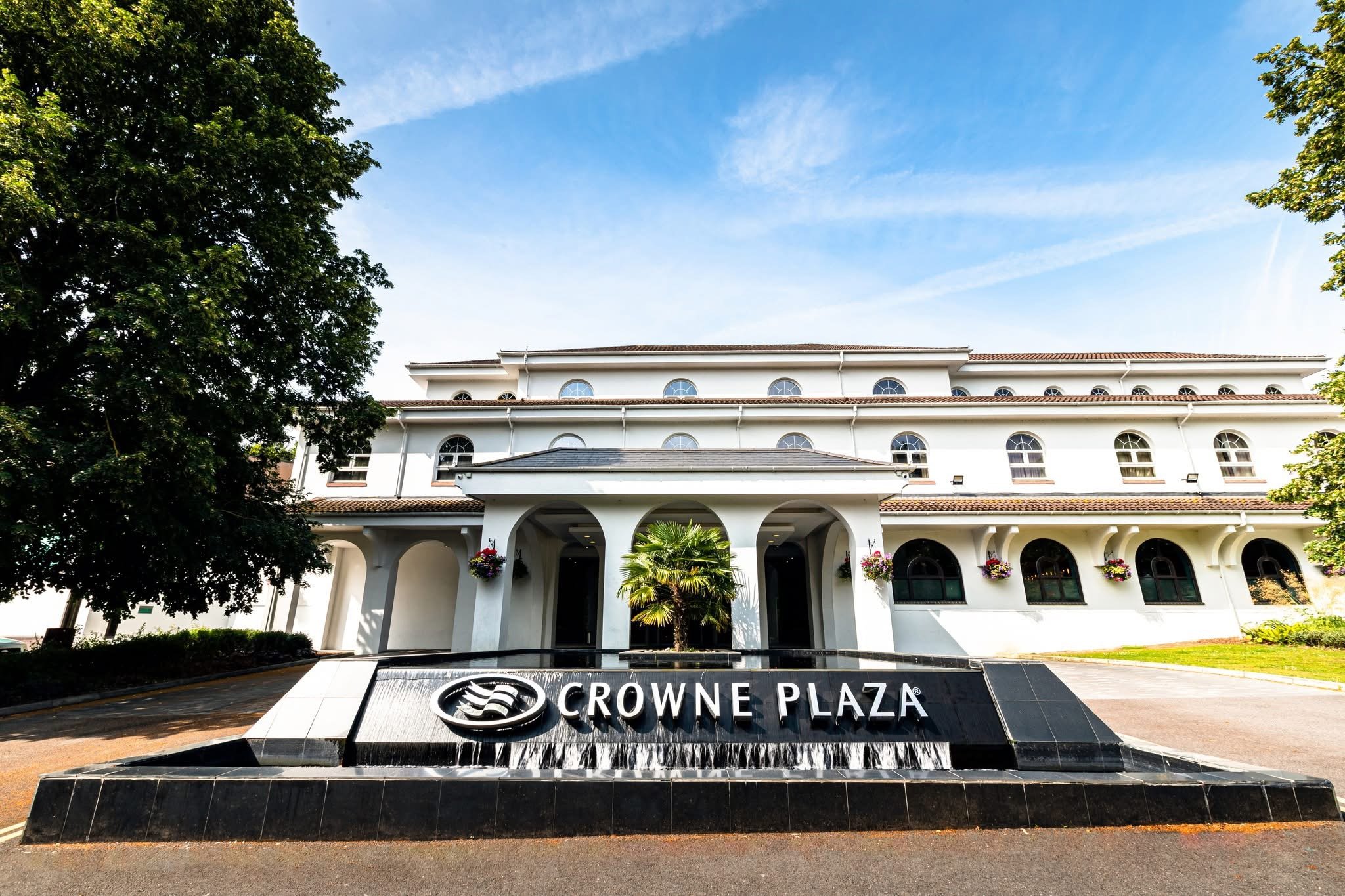 Front view of Crowne Plaza hotel with a water fountain and a black sign in the foreground, surrounded by greenery and trees, under a partly cloudy blue sky.