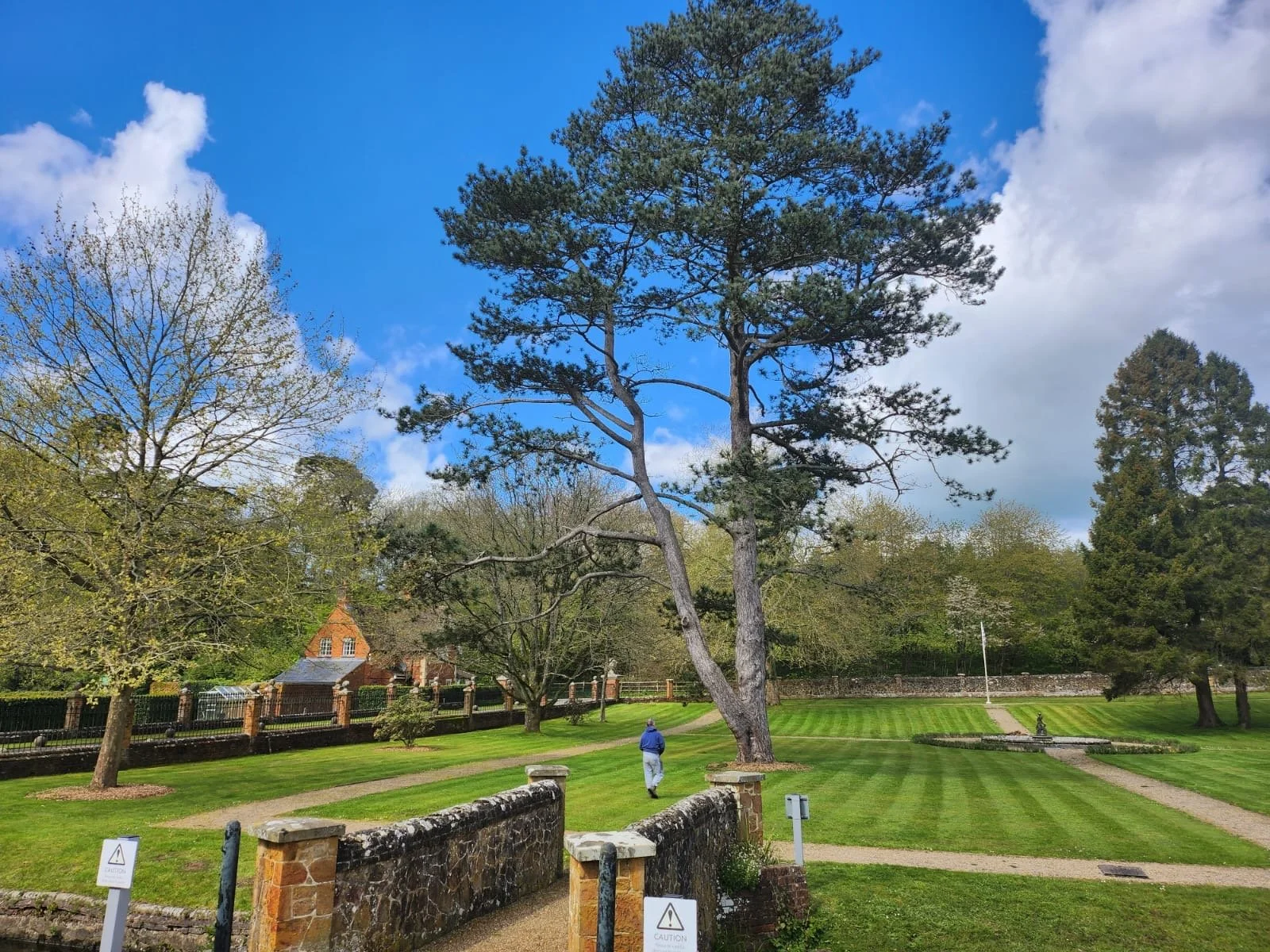 A park scene with trimmed green grass, tall trees, pathways, a person walking, and a partly cloudy sky.