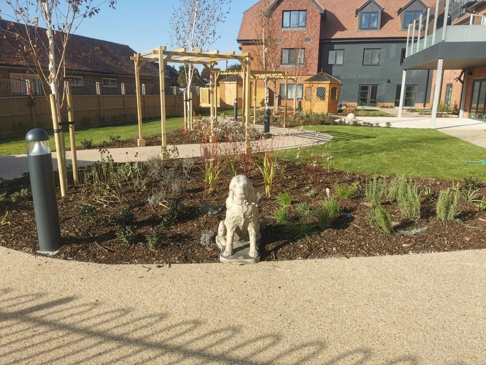 A landscaped garden with a lion statue in the foreground, a wooden pergola, a small wooden structure, and modern residential buildings in the background. The area is paved with concrete and features some grass and small plants.