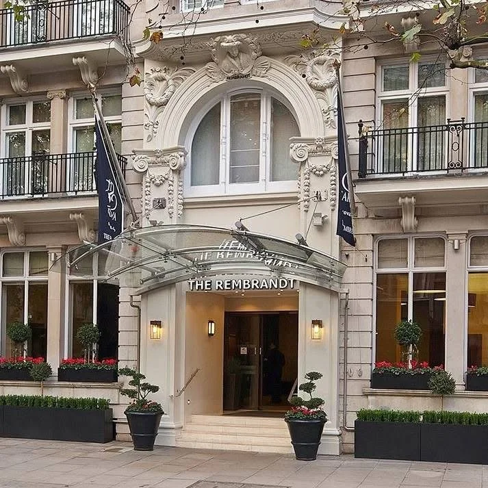 Hotel entrance with white ornate facade, glass canopy, and two flags. Potted plants and flowers decorate the entrance area.
