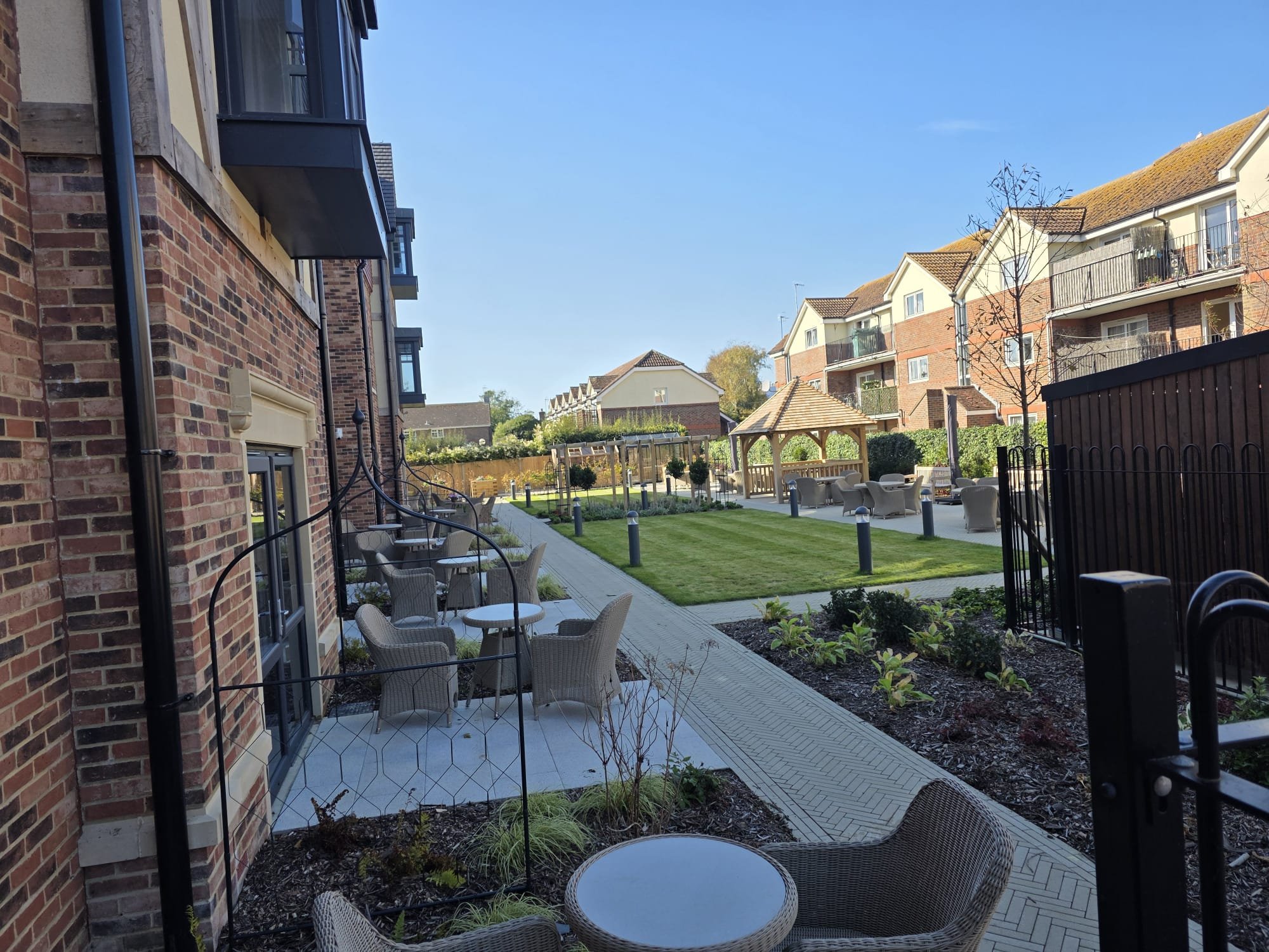 A peaceful outdoor courtyard with seating, a gazebo, and well-maintained lawn surrounded by apartment buildings on a clear sunny day.