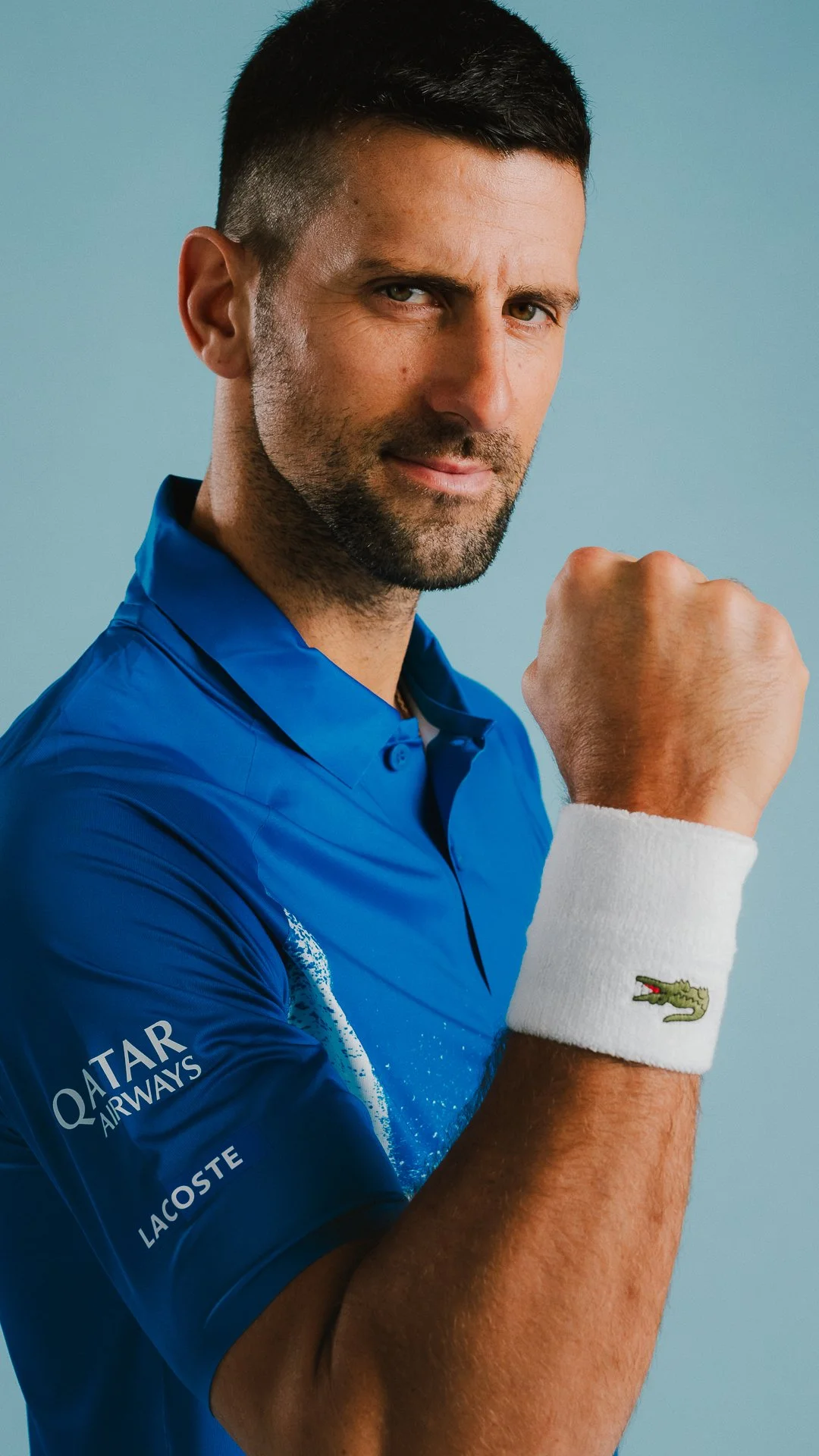A man wearing a blue shirt, with short dark hair and a beard, showing a confident expression and a clenched fist.