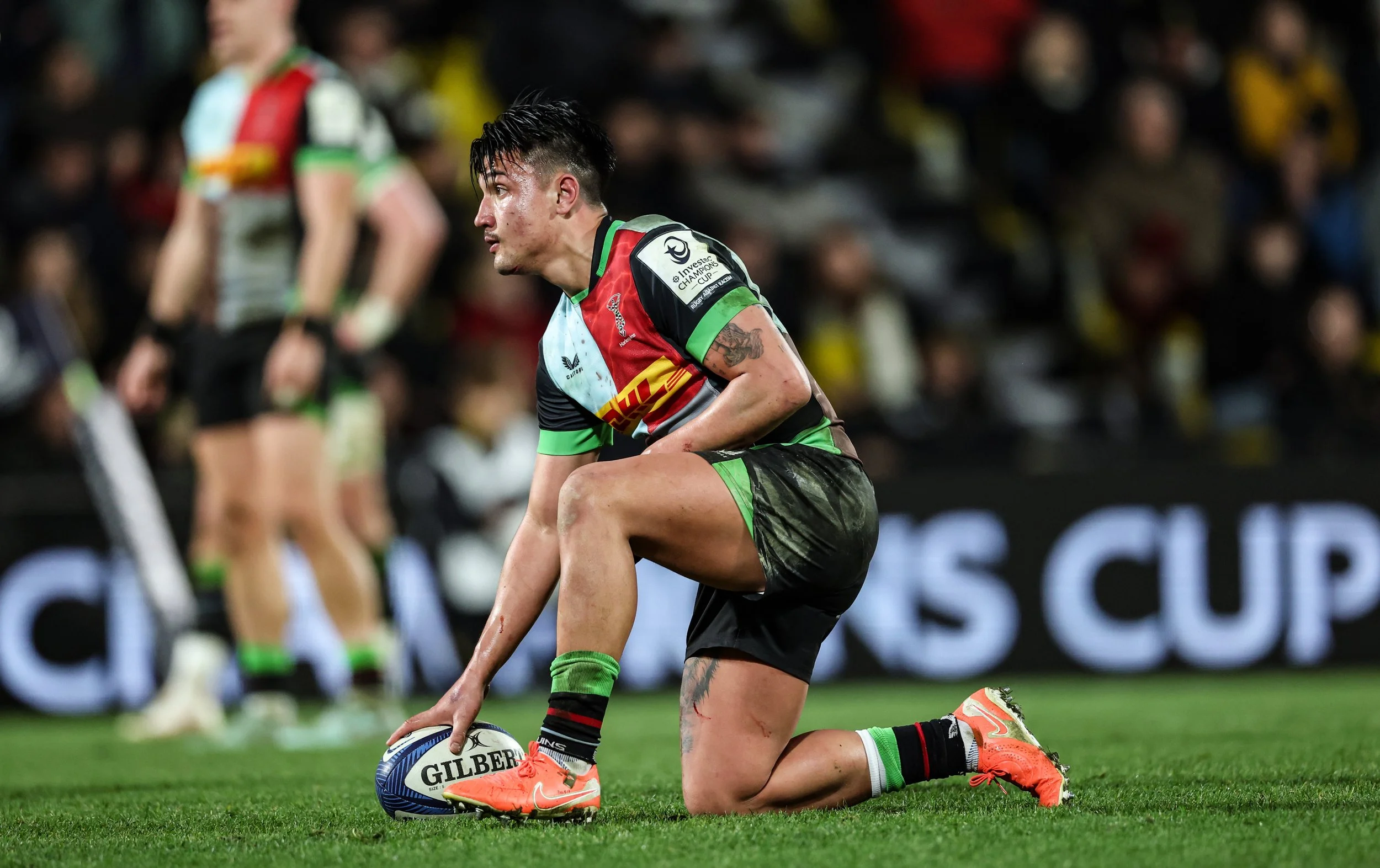 A rugby player in a colorful uniform kneeling on the field, holding a rugby ball, with a focused expression, during a match with a crowd in the background.
