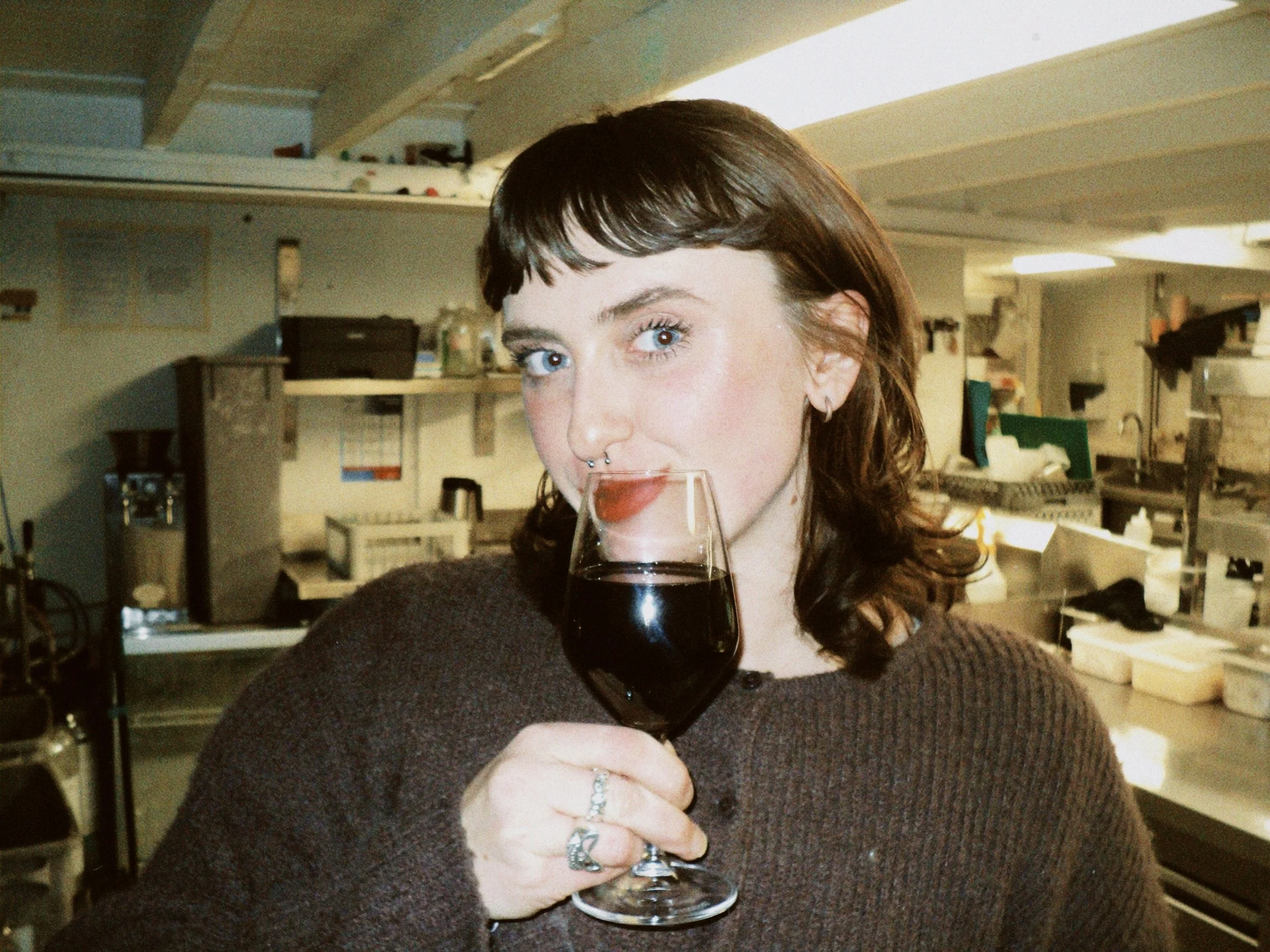 A woman with brown hair and blue eyes drinking red wine from a glass in a kitchen.
