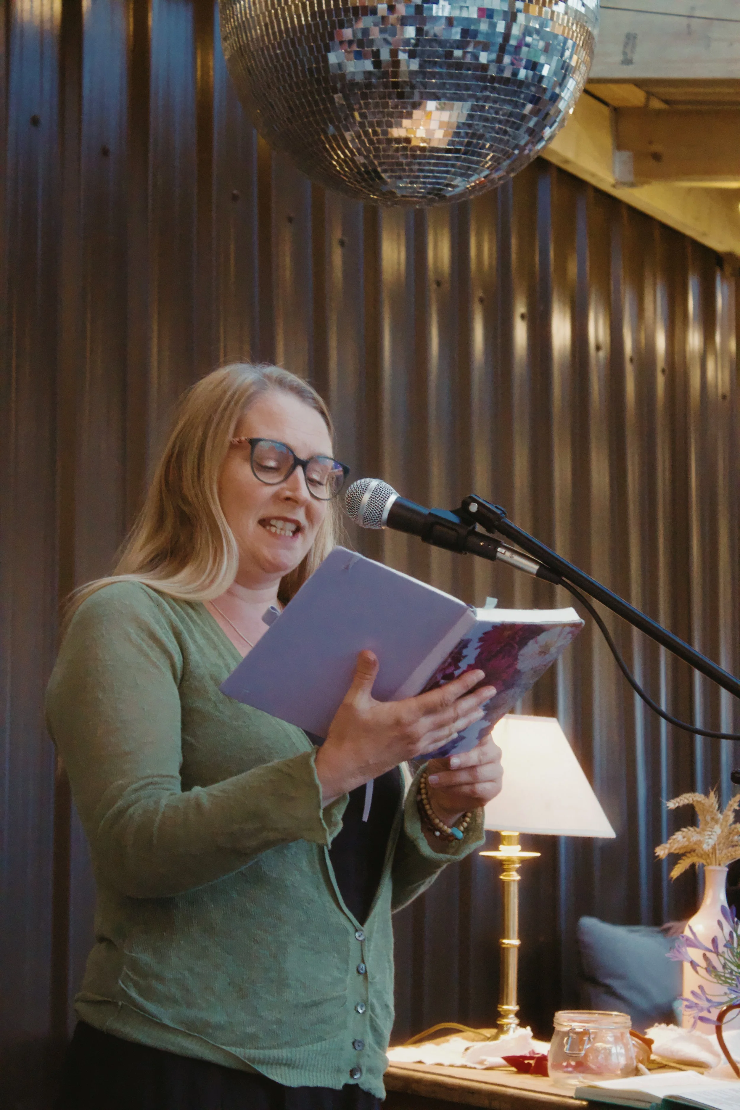 A woman with glasses and long blonde hair, wearing a green cardigan, is reading from a book at a microphone. The setting appears to be indoors with a wooden wall, a lamp, and decorative plants in the background. There is a large disco ball on the cei