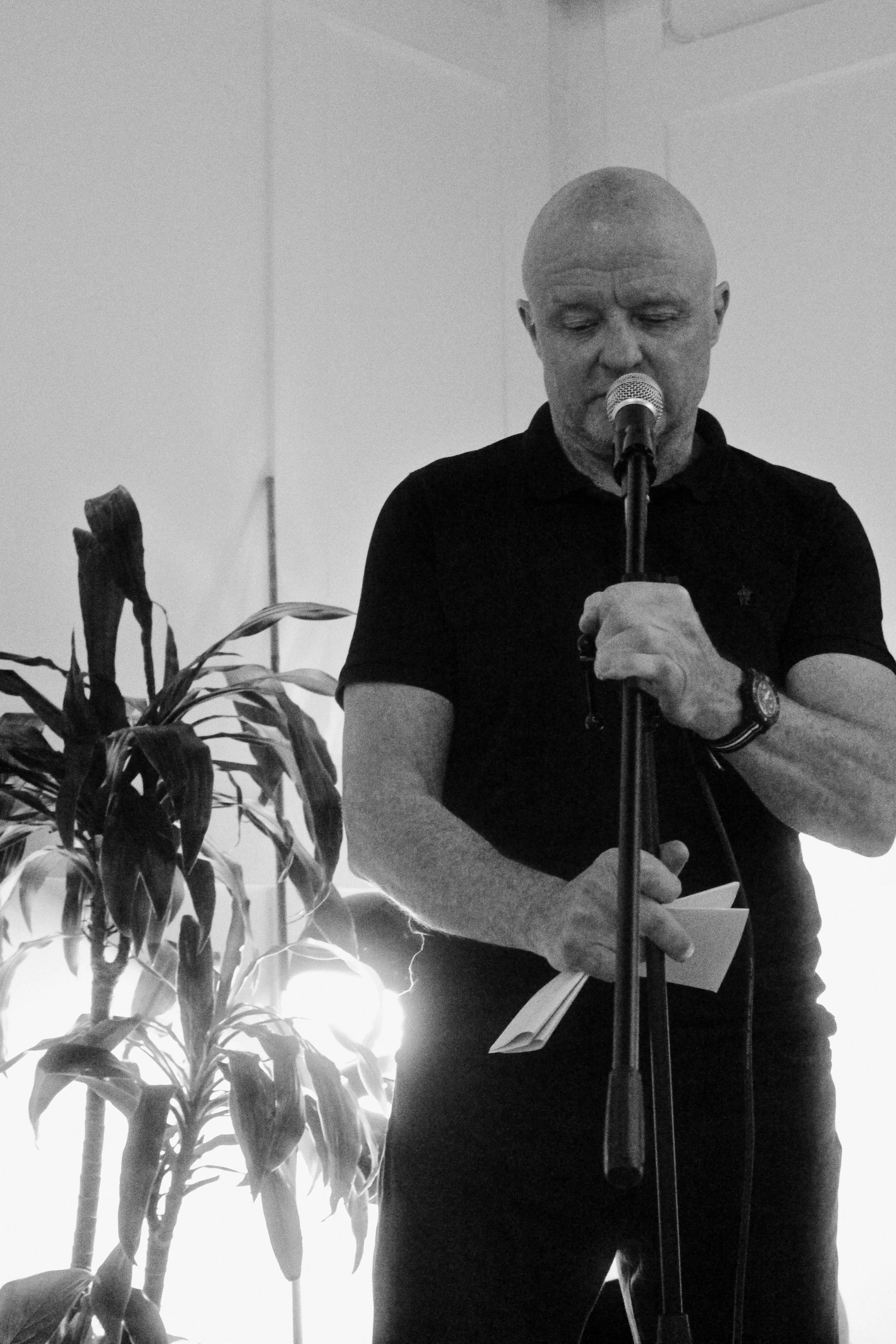 A man with a shaved head and a goatee is standing in front of a microphone, reading from a piece of paper. He is wearing a black polo shirt and a wristwatch. To his left, there is a potted plant with large, elongated leaves. The background is plain a