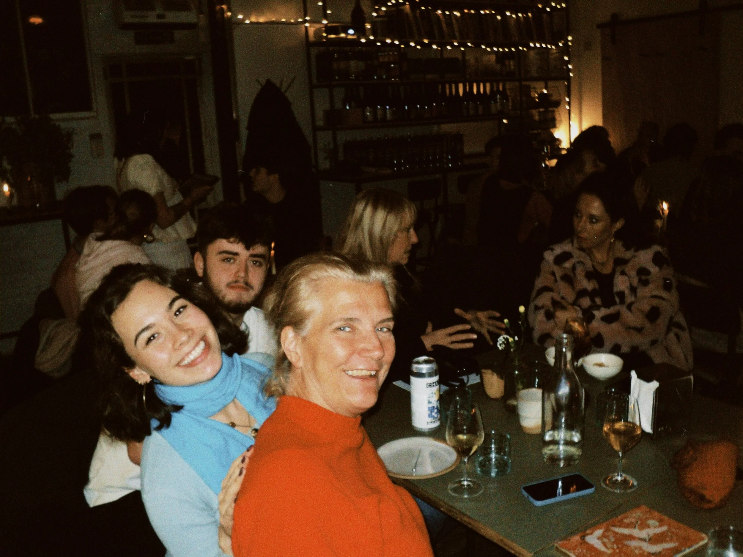 A group of people gathered at a dinner event, sitting at a table with wine glasses and bottles, in a dimly lit room with candles and shelves in the background.