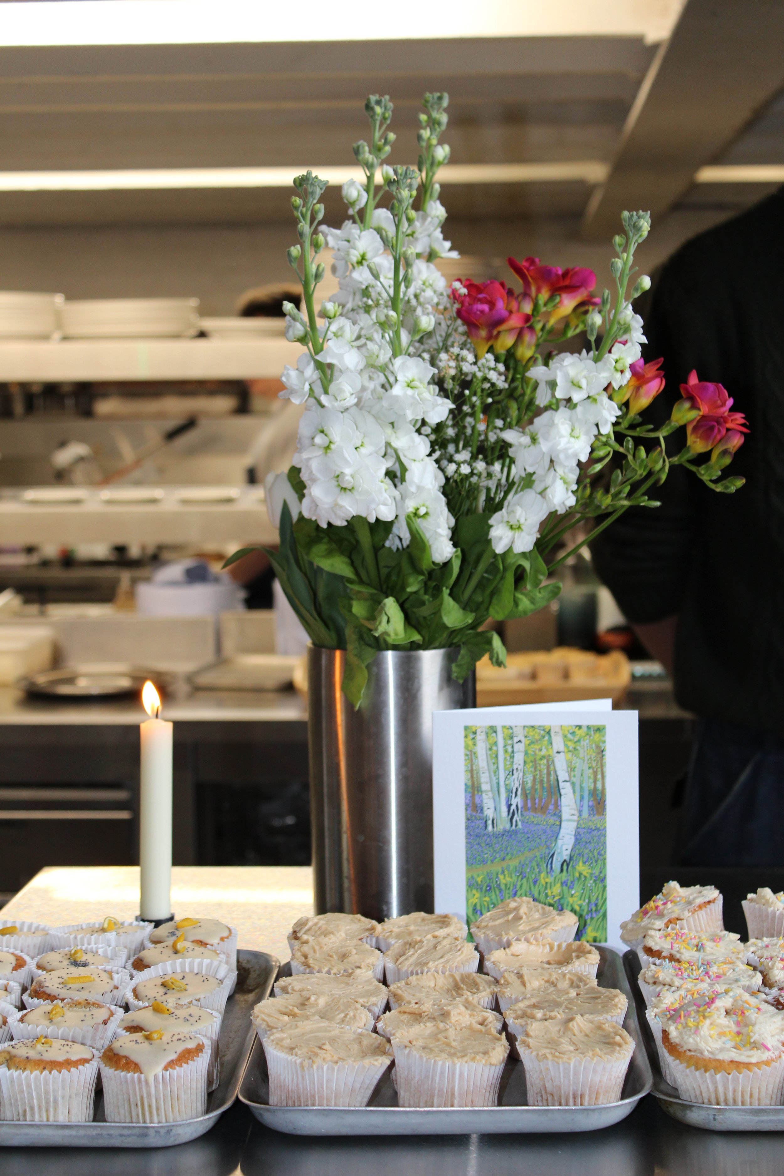 Tray of cupcakes with white frosting, some decorated with sprinkles or small yellow decorations, and a candle next to them, in a bakery or cafe setting with flowers and a painting of a forest scene in the background.