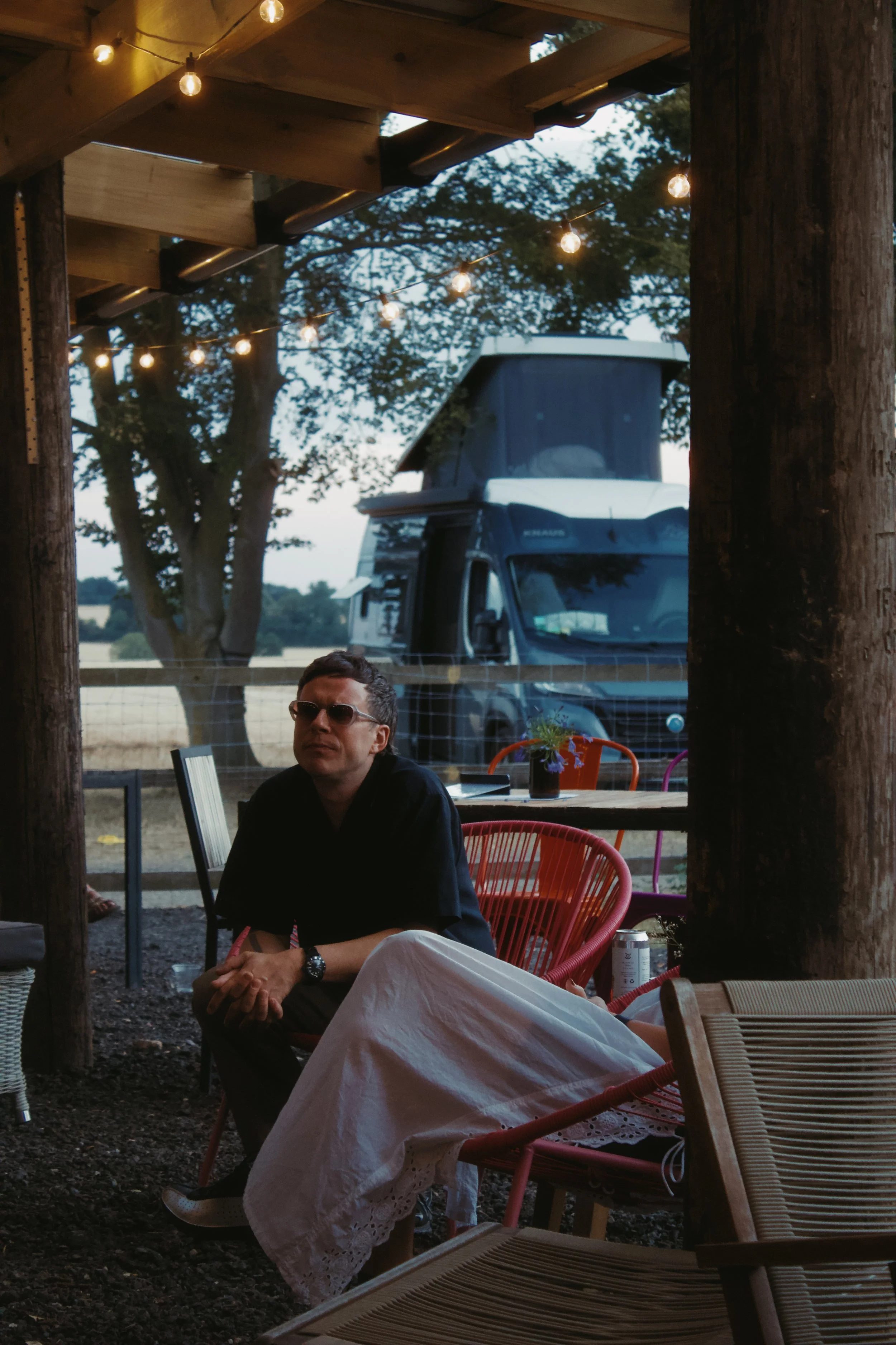 A man with sunglasses sitting outdoors at a rustic table with string lights overhead, trees and a camper van in the background, and another person partially visible lying on the ground.
