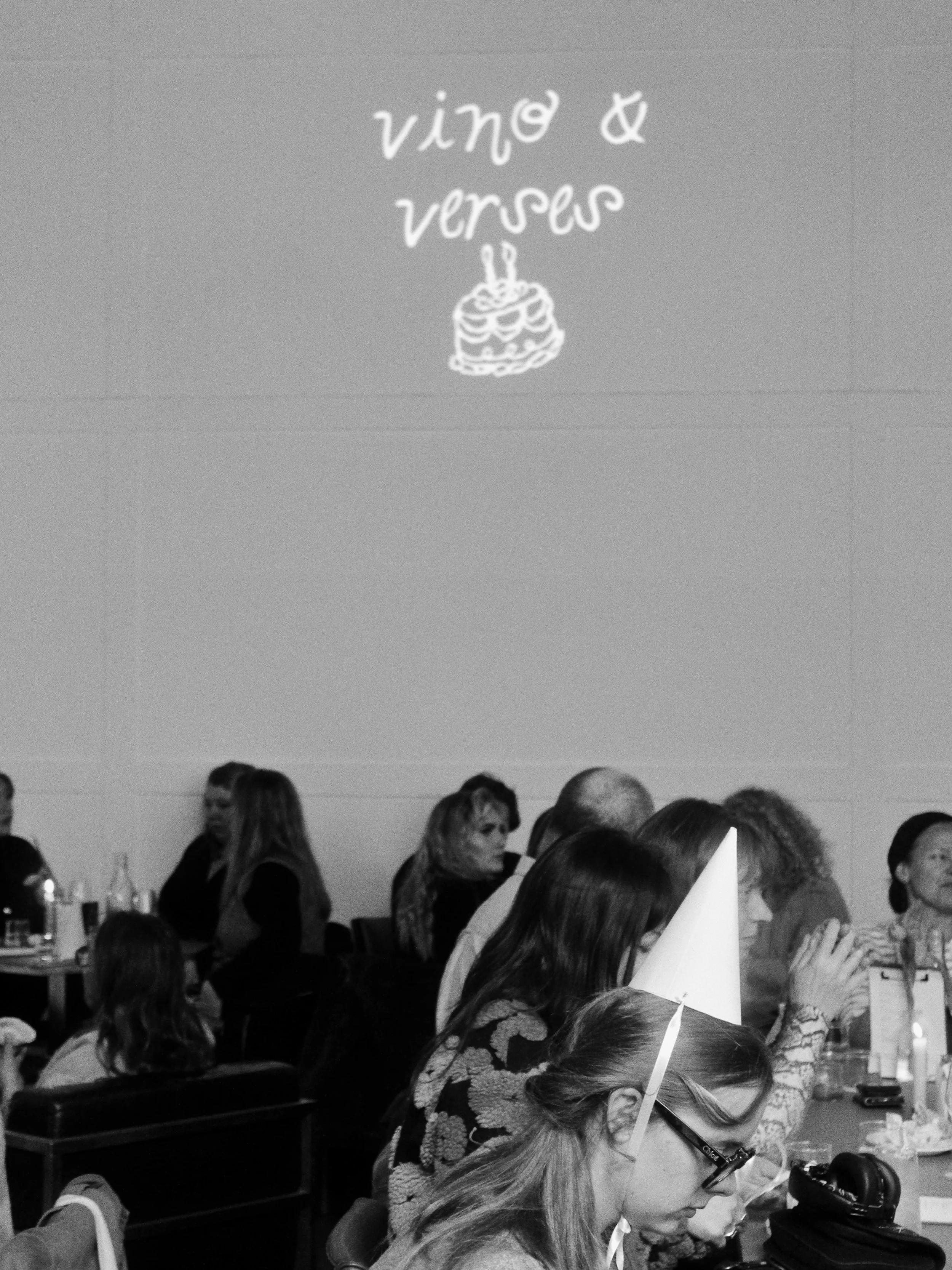 A group of people sitting around tables in a room decorated for a celebration, with a sign on the wall that reads 'vino & veritas' and a hand-drawn cake illustration. Some are eating or talking, and there is a woman with glasses in the foreground loo