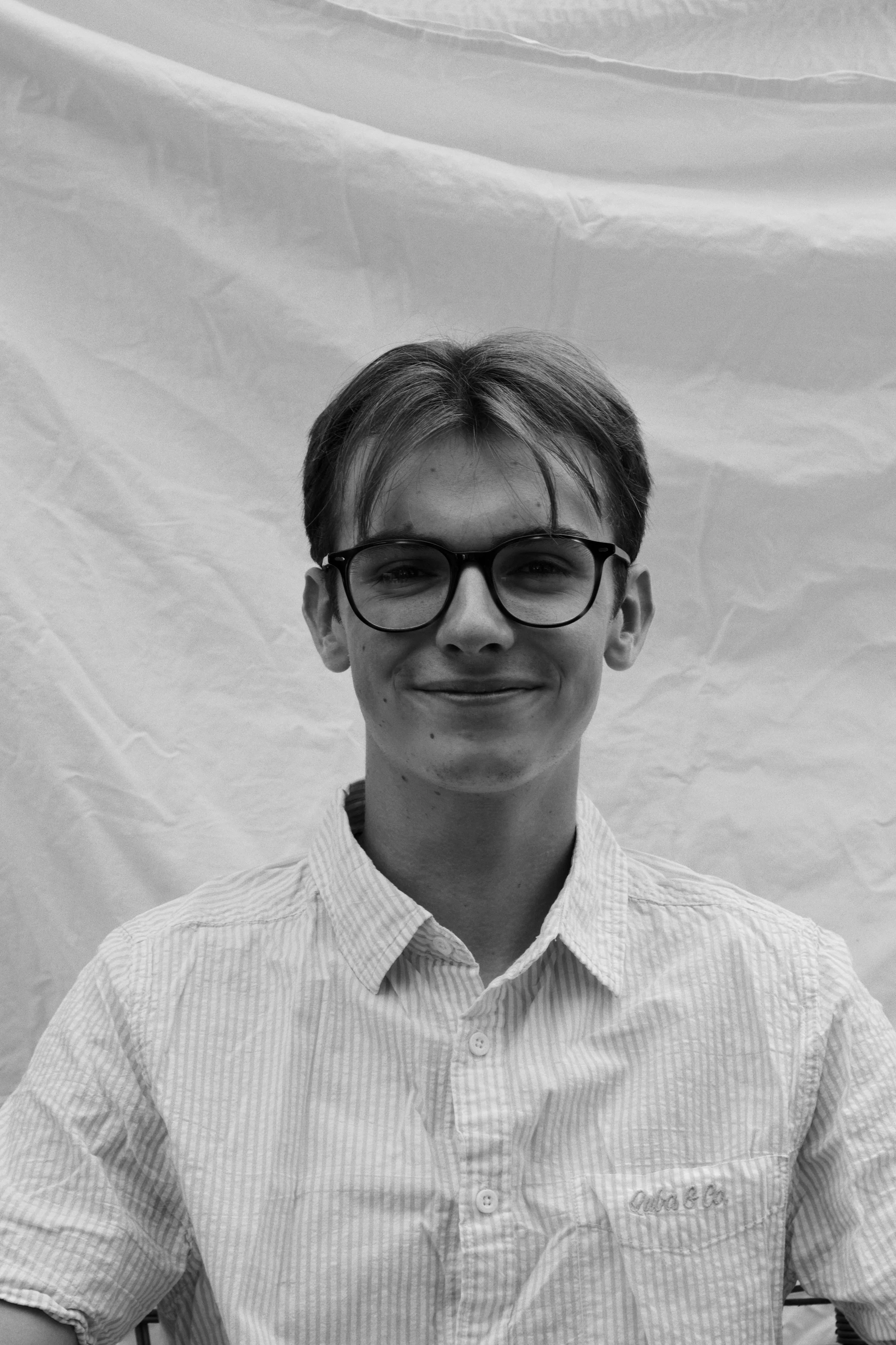 Black and white photo of a smiling young man wearing glasses and a striped collared shirt, standing against a wrinkled background.