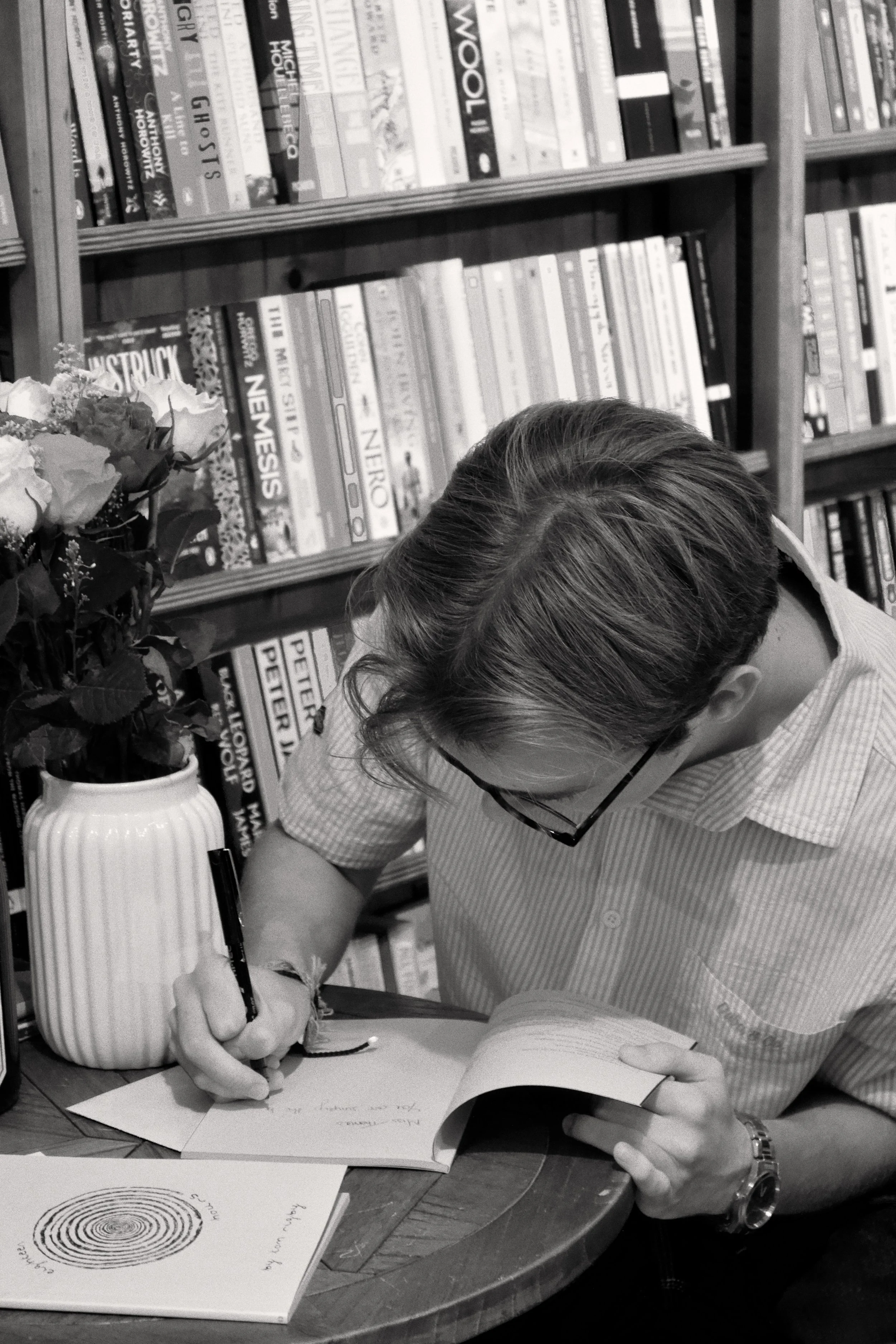 A person sitting at a table, writing in a notebook with a pen. They are wearing glasses and a striped shirt, with bookshelves filled with books in the background. There is a vase with flowers on the table and some open books.