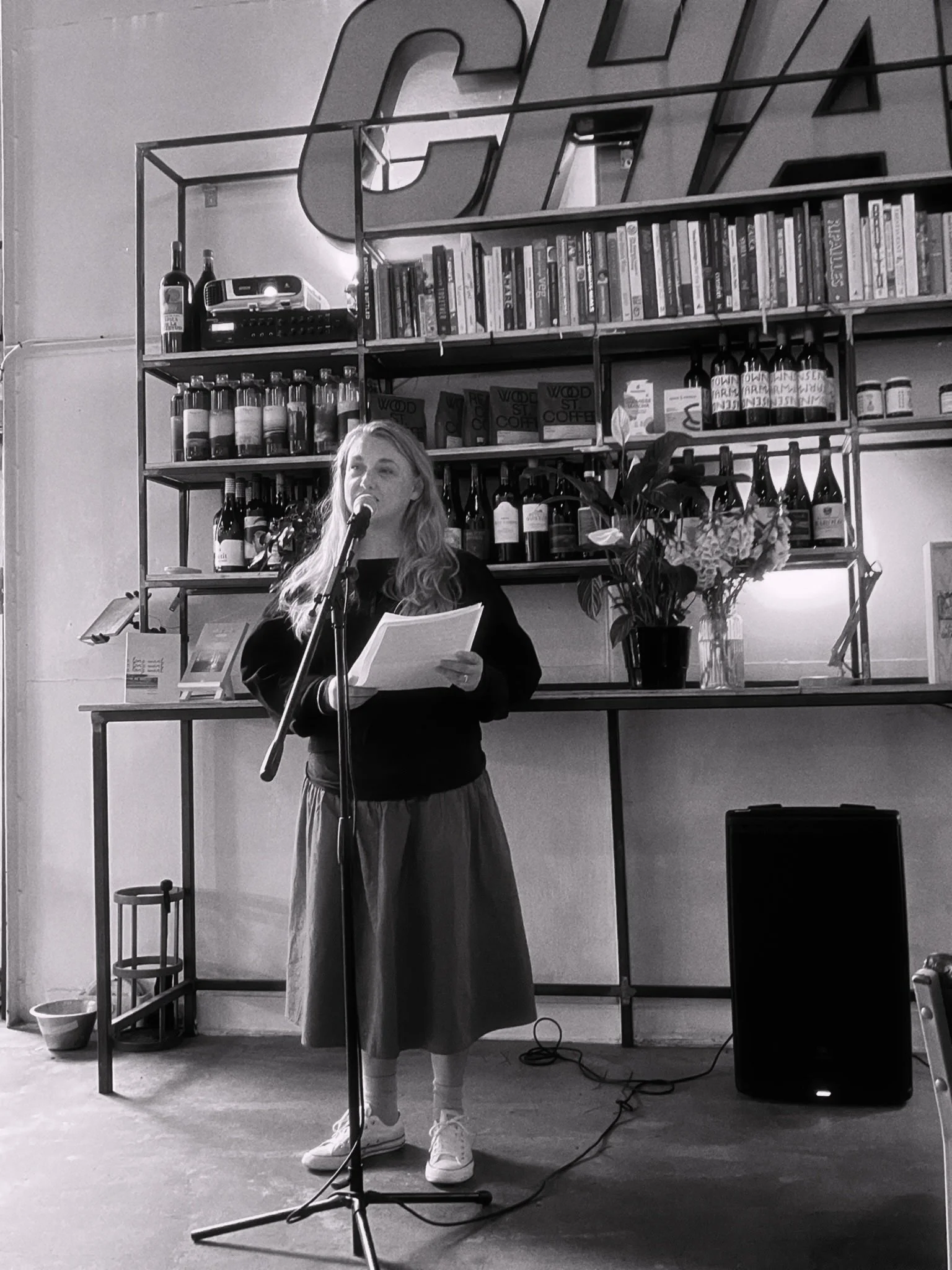 A woman standing with a microphone, reading from a paper, on a stage with a bookshelf behind her, decorated with bottles, books, and plants.
