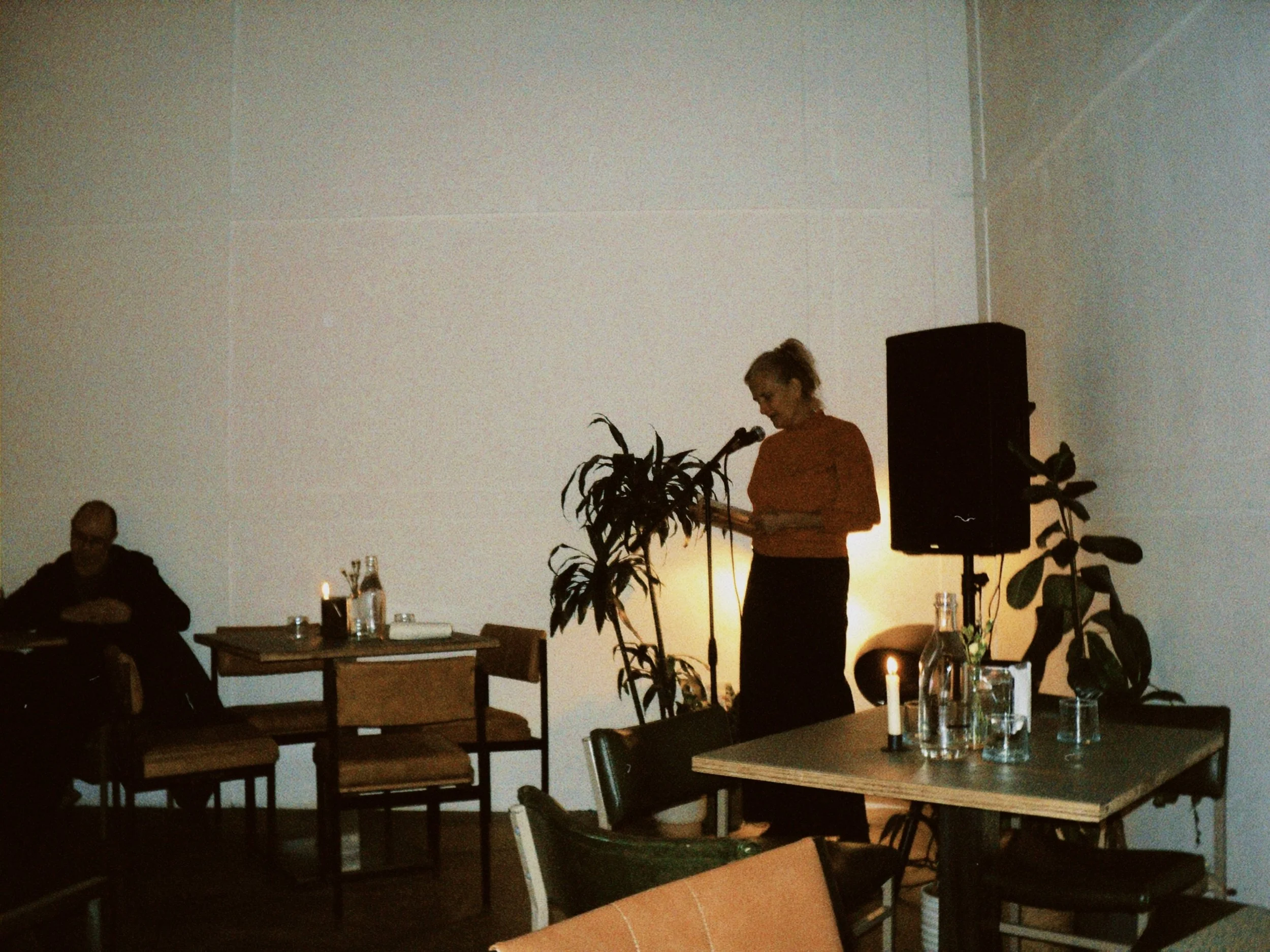 A woman stands at a microphone, speaking during an indoor event, with a large speaker and potted plants nearby. There are candles and drinking glasses on the tables, with a man sitting at a table to the left.
