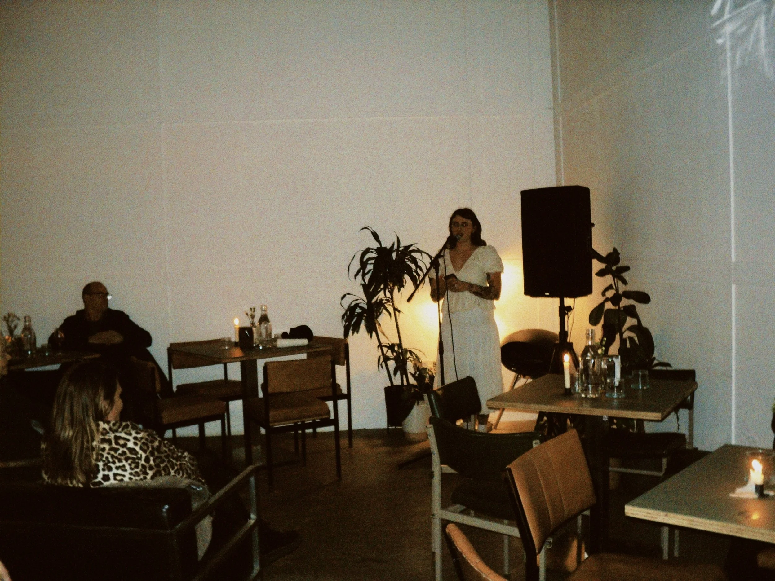 A woman in a white dress speaking into a microphone at an indoor event with seated audience members and tables with drinks and candles.