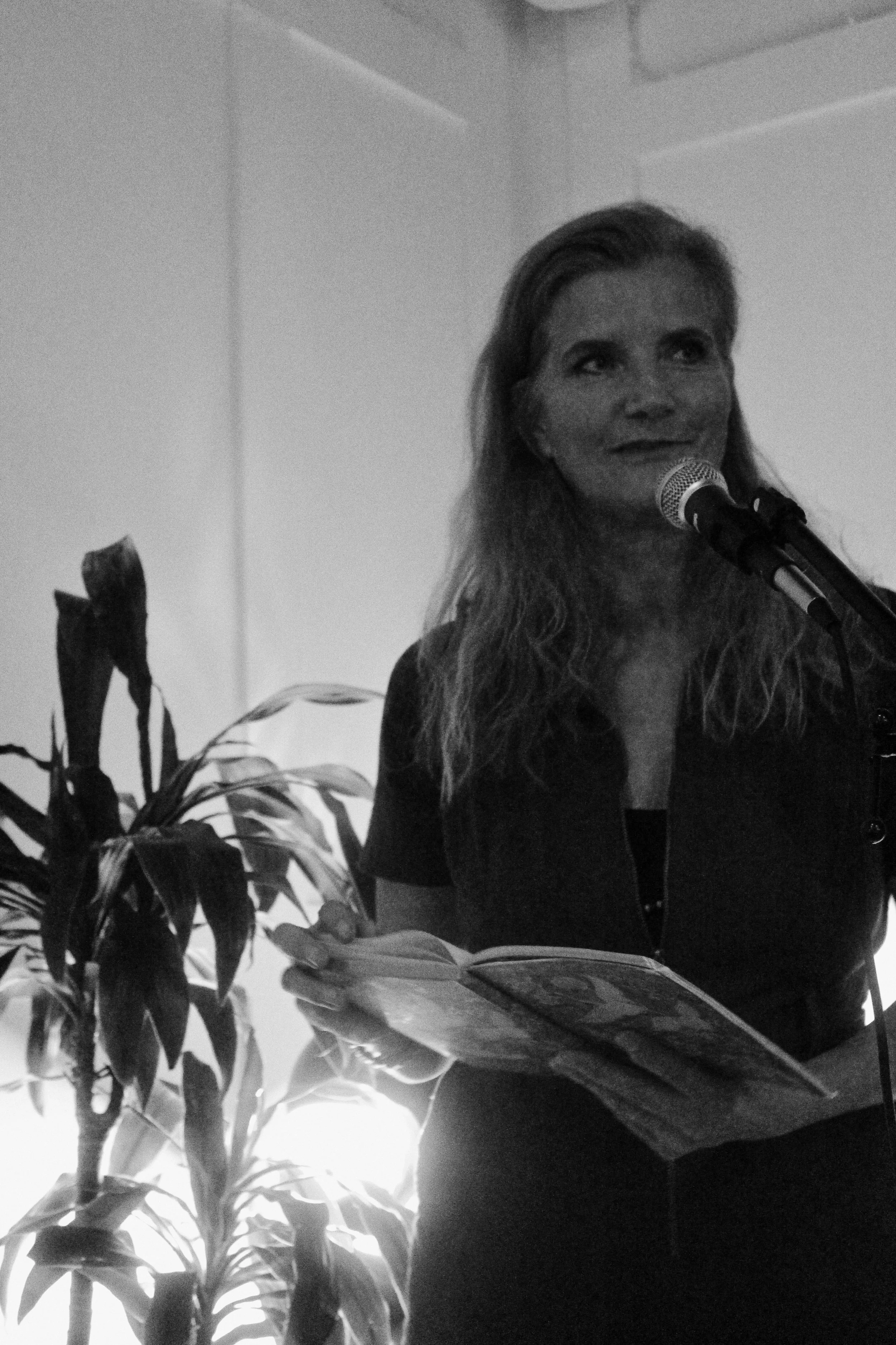 Woman speaking into a microphone at a table, with papers and books in front, and a potted plant in the background.