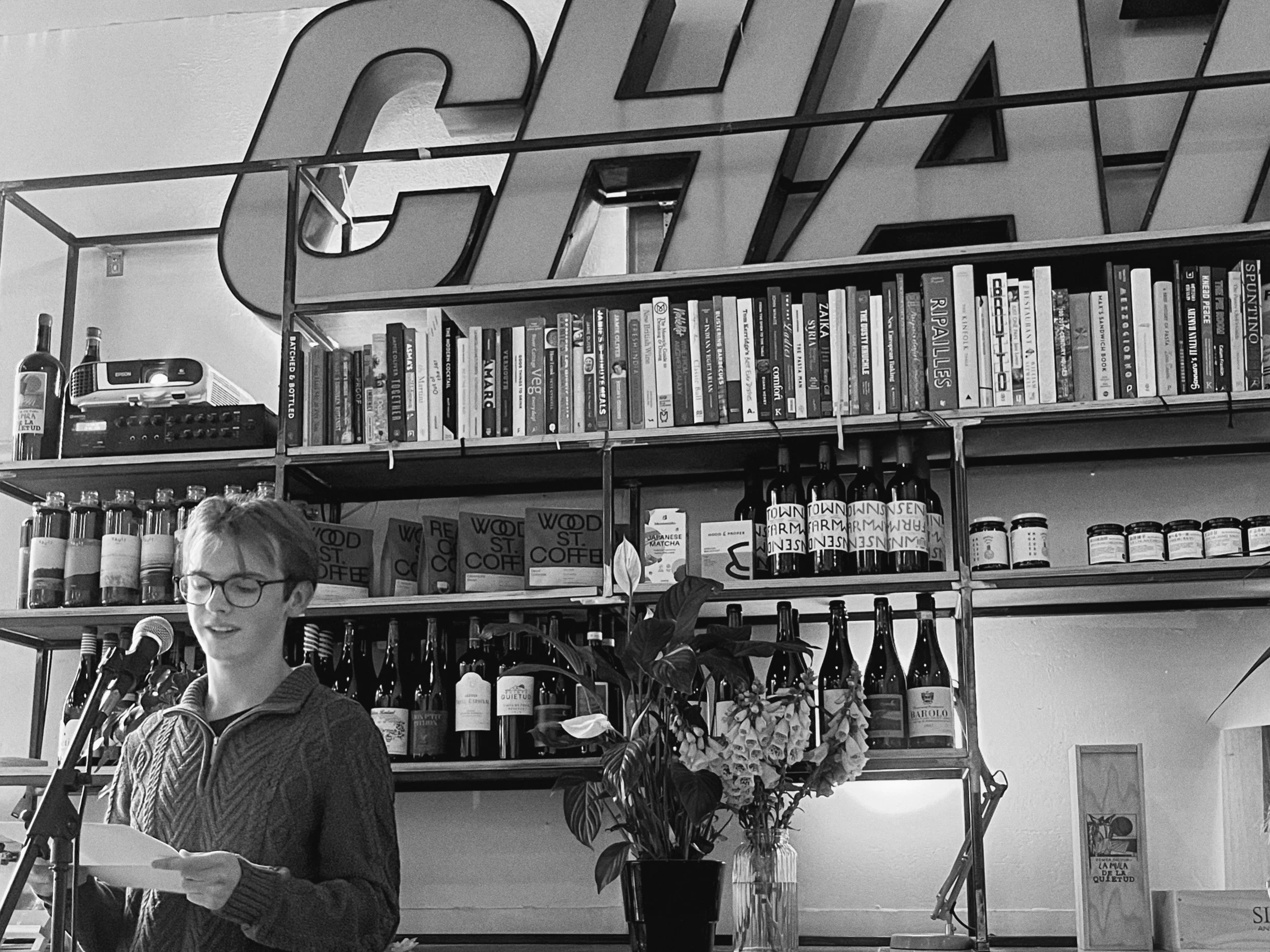 A woman with glasses and short hair, wearing a patterned sweater, speaking into a microphone at a bookstore or cafe. Behind her is a bookshelf filled with various books and bottles, and a decorative floral arrangement.