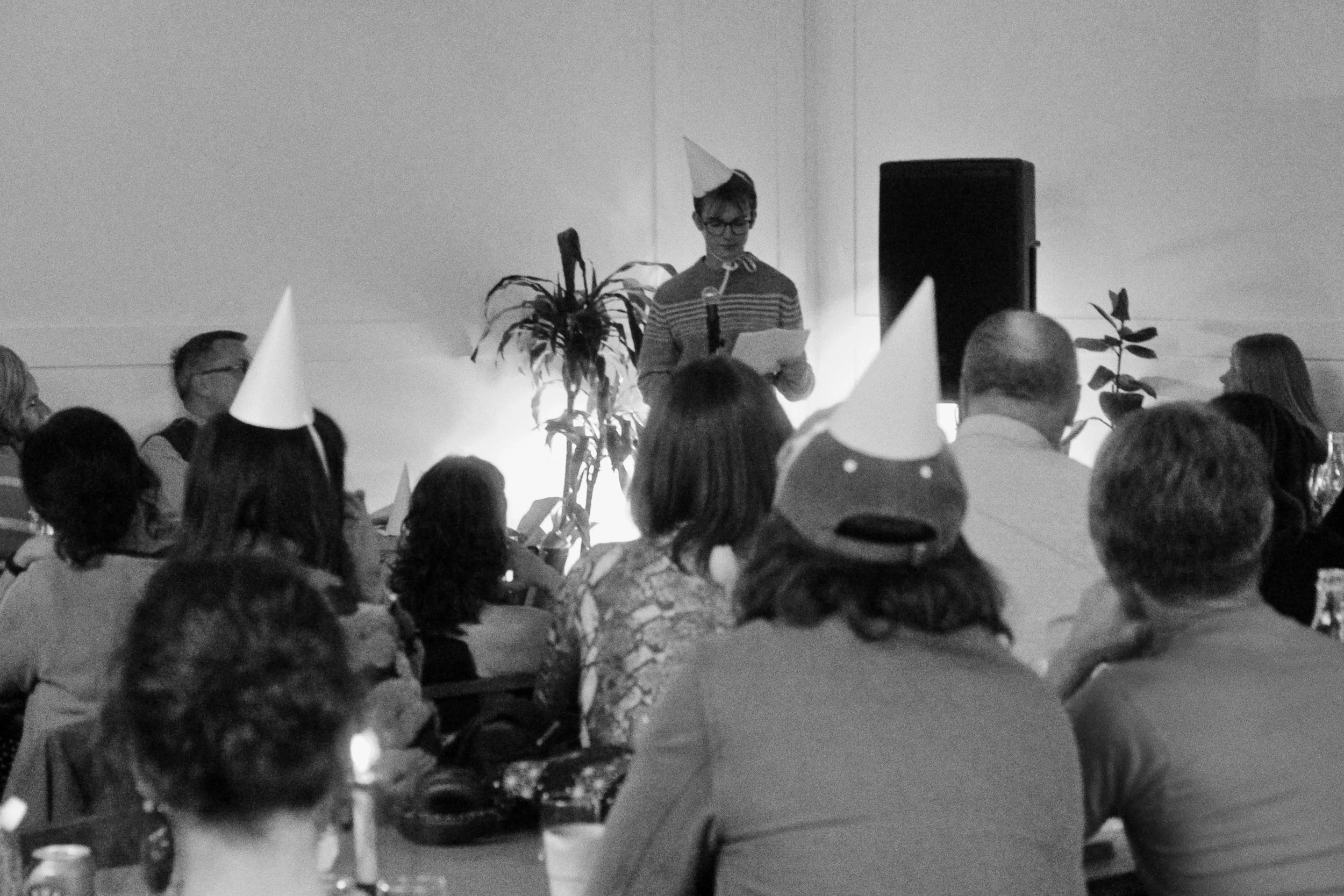 A person wearing glasses and a party hat reading from a paper at a celebration, with guests wearing party hats seated at tables.