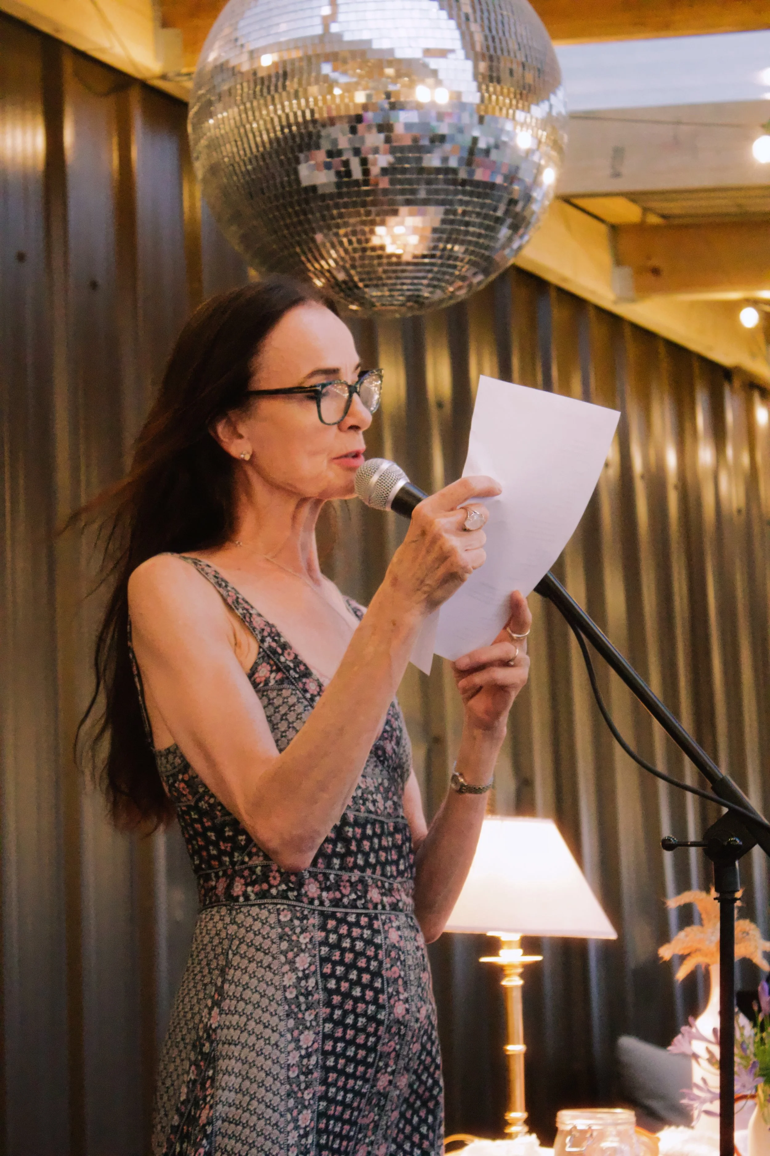 A woman with long dark hair, glasses, and jewelry reading a paper into a microphone at a decorated indoor event with a disco ball overhead and warm lighting.