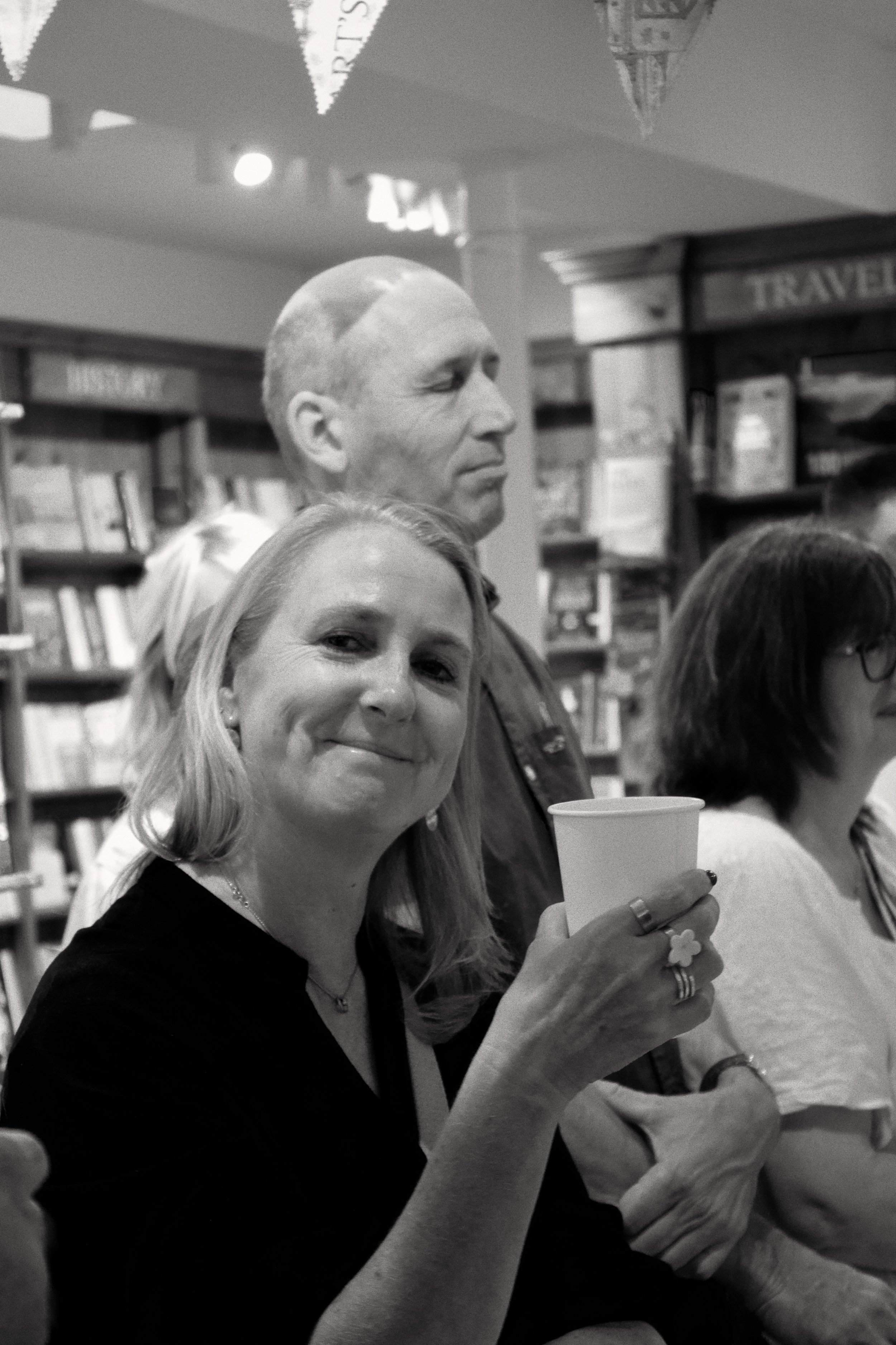 Black and white photo of several people, including a woman in the foreground holding a cup, smiling at the camera. Behind her, a man with short hair and a serious expression is visible, with bookshelves and decorations in the background, suggesting a