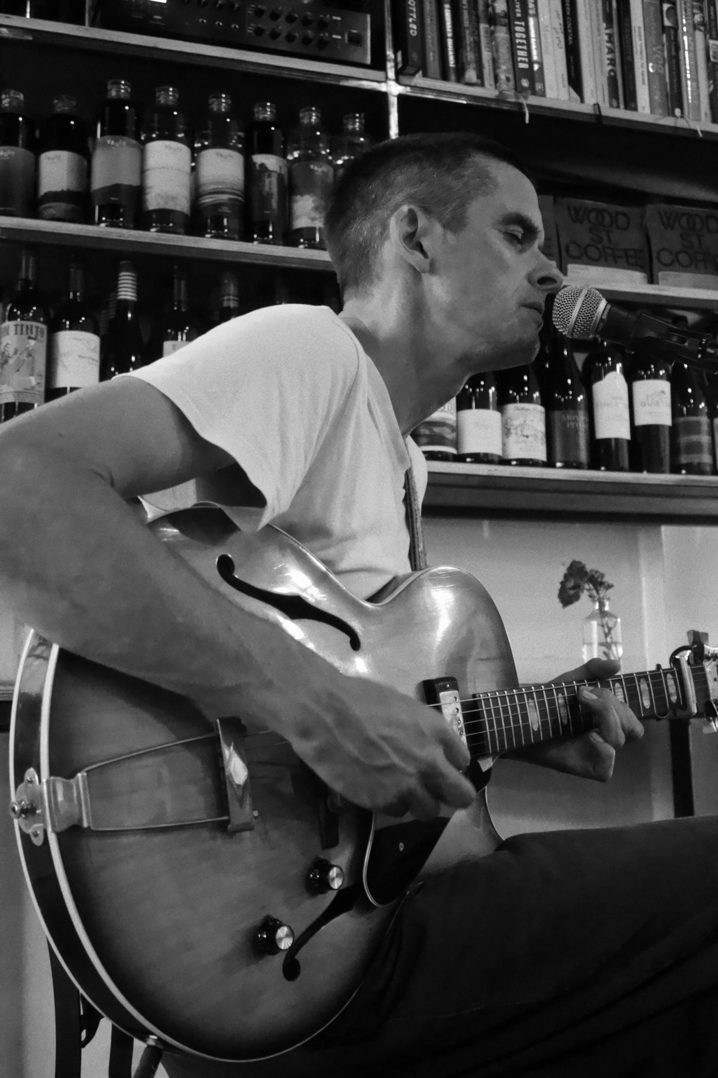 A young man with short hair is playing an acoustic guitar and singing into a microphone in a room with shelves of books and bottles in the background.
