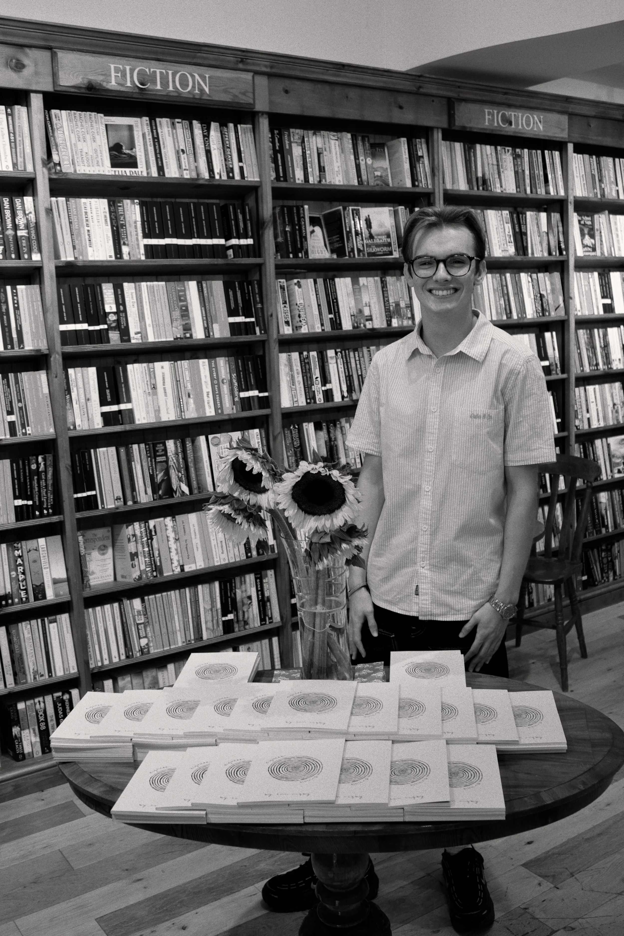 A smiling young man with glasses stands in front of bookshelves labeled 'Fiction' in a bookstore. On the table in front of him, there are numerous copies of a book with a concentric circle design on the cover. A bouquet of sunflowers is next to the b