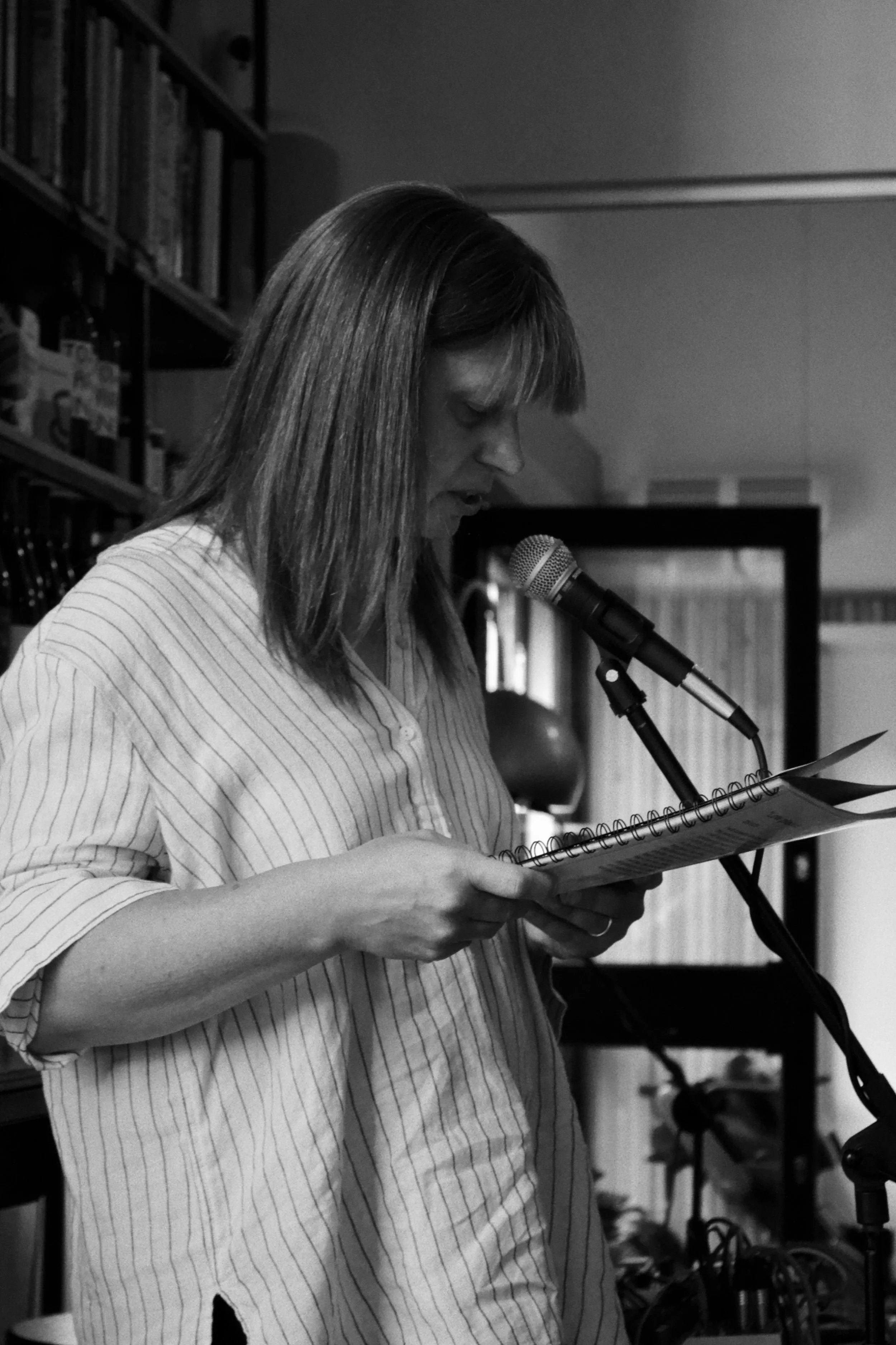A woman in a striped shirt reading from a paper into a microphone in a room with bookshelves.